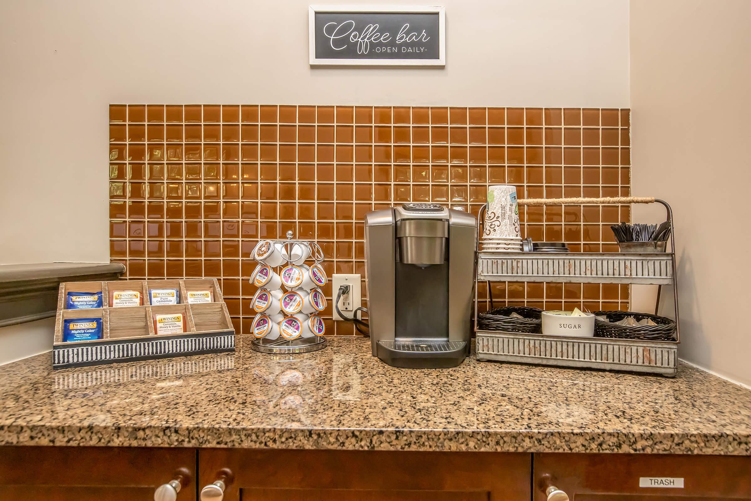 A cozy coffee bar setup featuring a coffee machine on a granite countertop. To the left, there are assorted tea bags and coffee pods arranged neatly in a circular holder. To the right, there are utensils in a container, alongside a sign reading "STIR." The backsplash is tiled in warm brown tones, creating an inviting atmosphere.