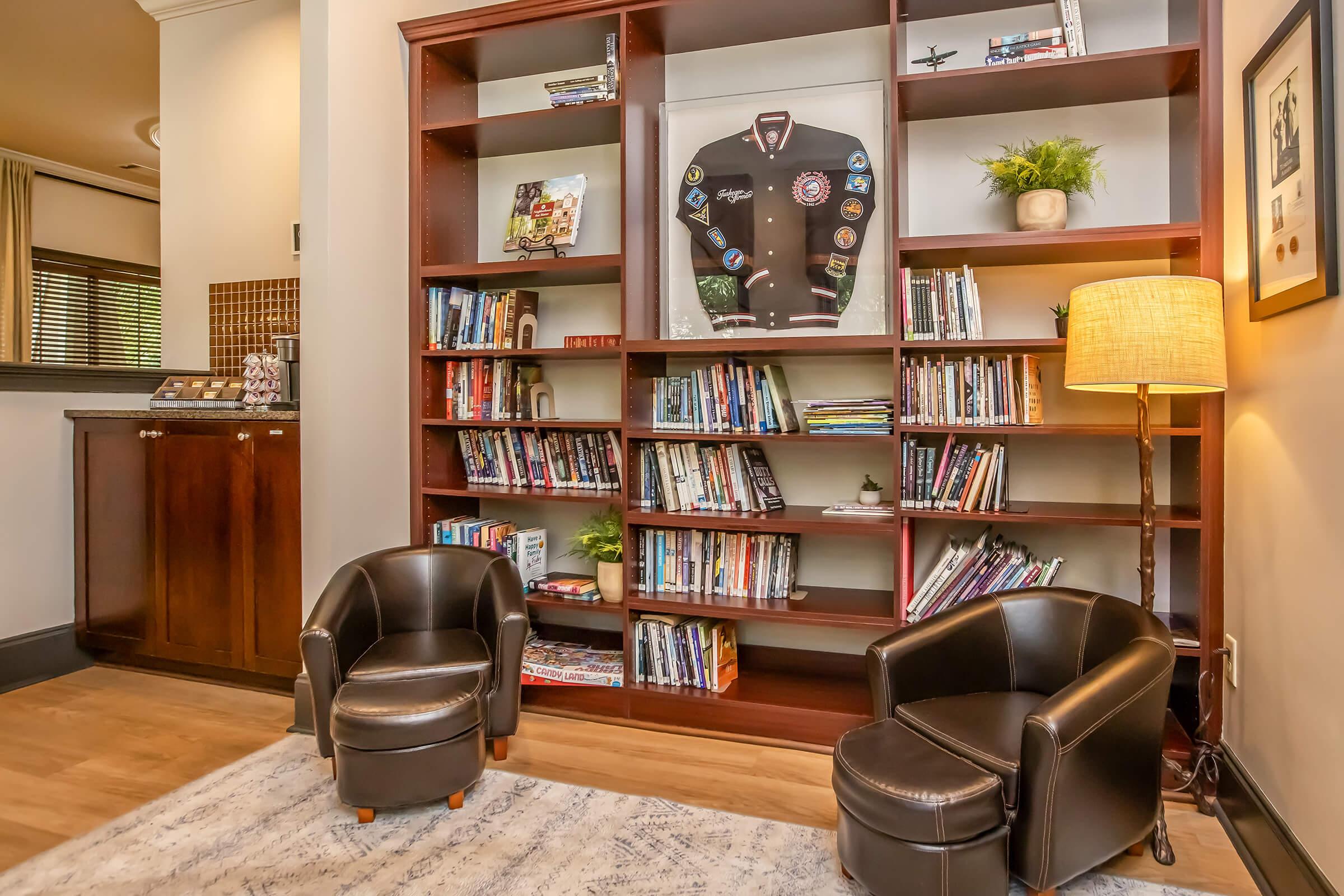 Cozy reading nook featuring two black armchairs in front of a tall bookshelf filled with books. A framed varsity jacket hangs on the wall, and a small potted plant is placed on a shelf. Warm lighting from a nearby lamp adds to the inviting atmosphere. A wooden cabinet is visible in the background.