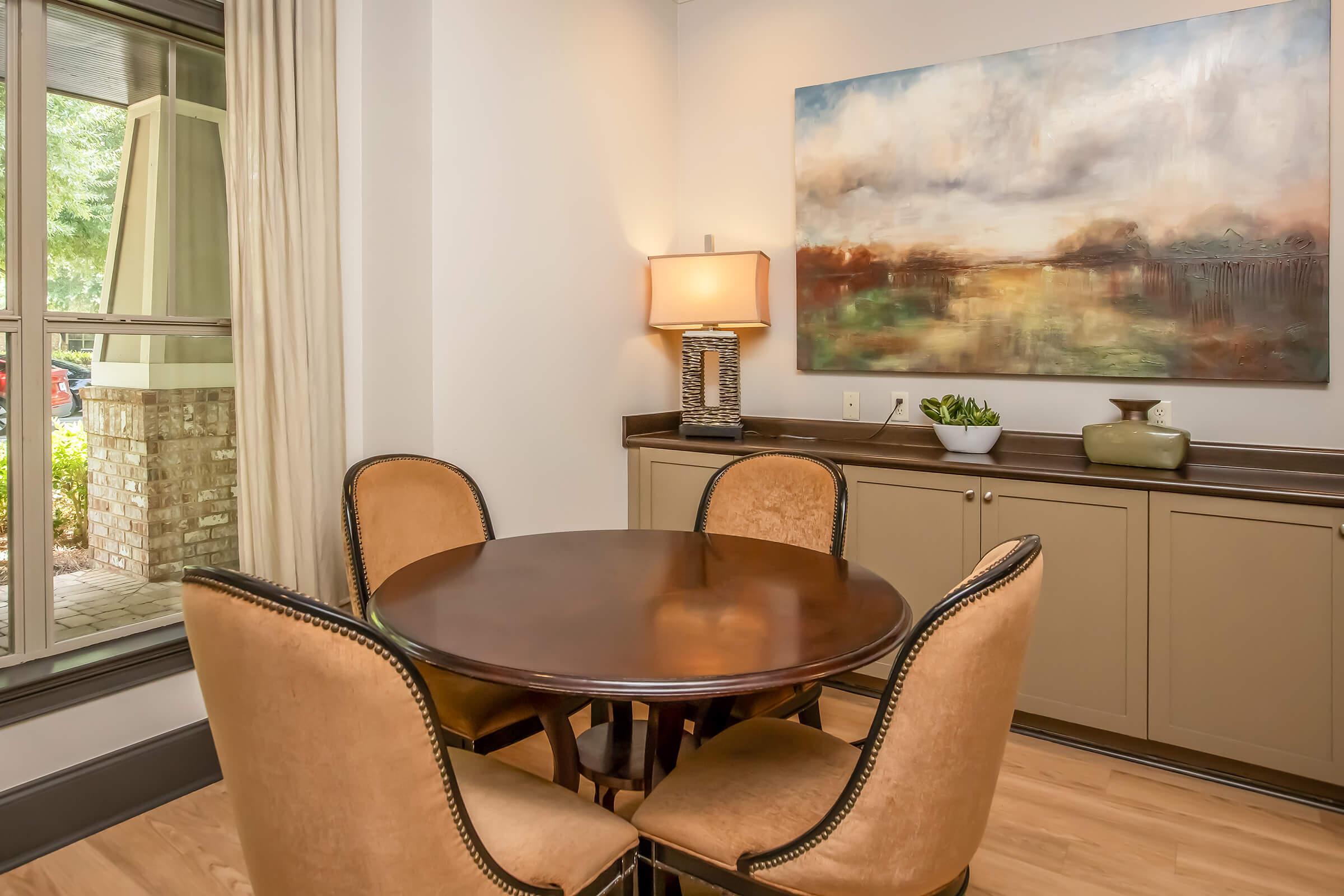 A cozy dining area featuring a round wooden table surrounded by four upholstered chairs. There is a lamp on a side table, and a large abstract painting is hanging on the wall. A potted plant and decorative items sit on a low cabinet. Natural light flows in through a nearby window.
