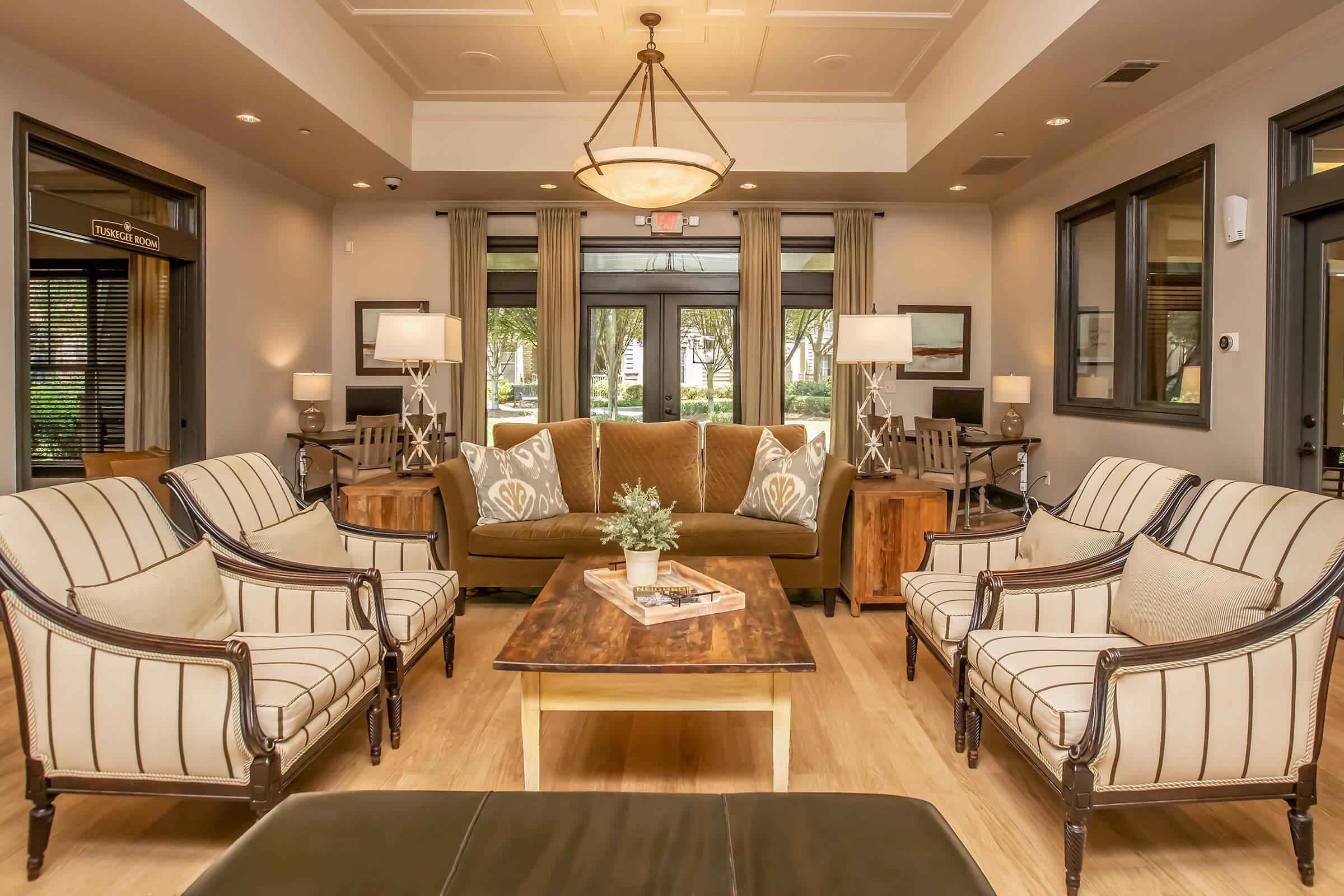 Cozy and stylish lounge area featuring a brown sofa with decorative pillows, two elegant armchairs, a wooden coffee table with a decorative plant, and a bright atmosphere with large windows. Soft lighting from table lamps enhances the inviting ambiance.