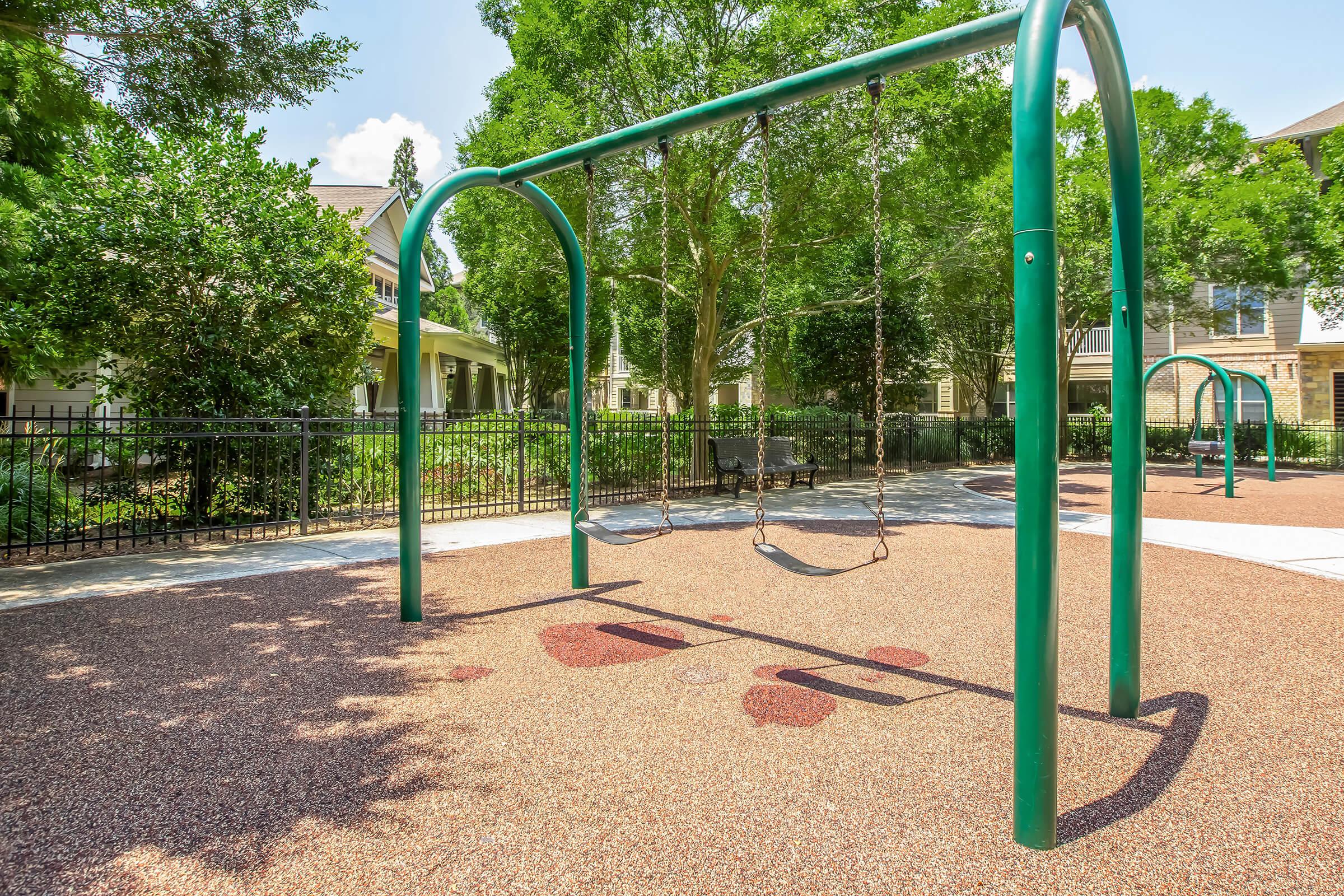 Empty swings on a playground surrounded by trees and green grass. The ground is covered in a textured rubber surface, and a shaded bench is visible in the background. The area is well-maintained and inviting for children.