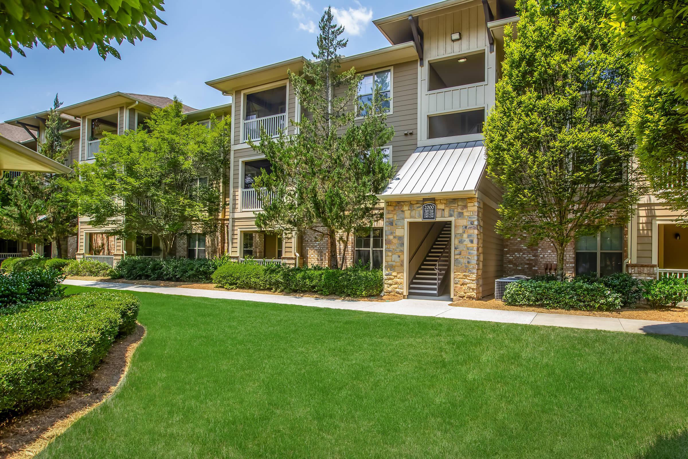 Three-story apartment building surrounded by lush green landscaping, shrubs, and small trees. The building features a mix of stone and siding exterior, with balconies visible on the upper floors. A pathway leads to a staircase entering the building, set against a bright blue sky.