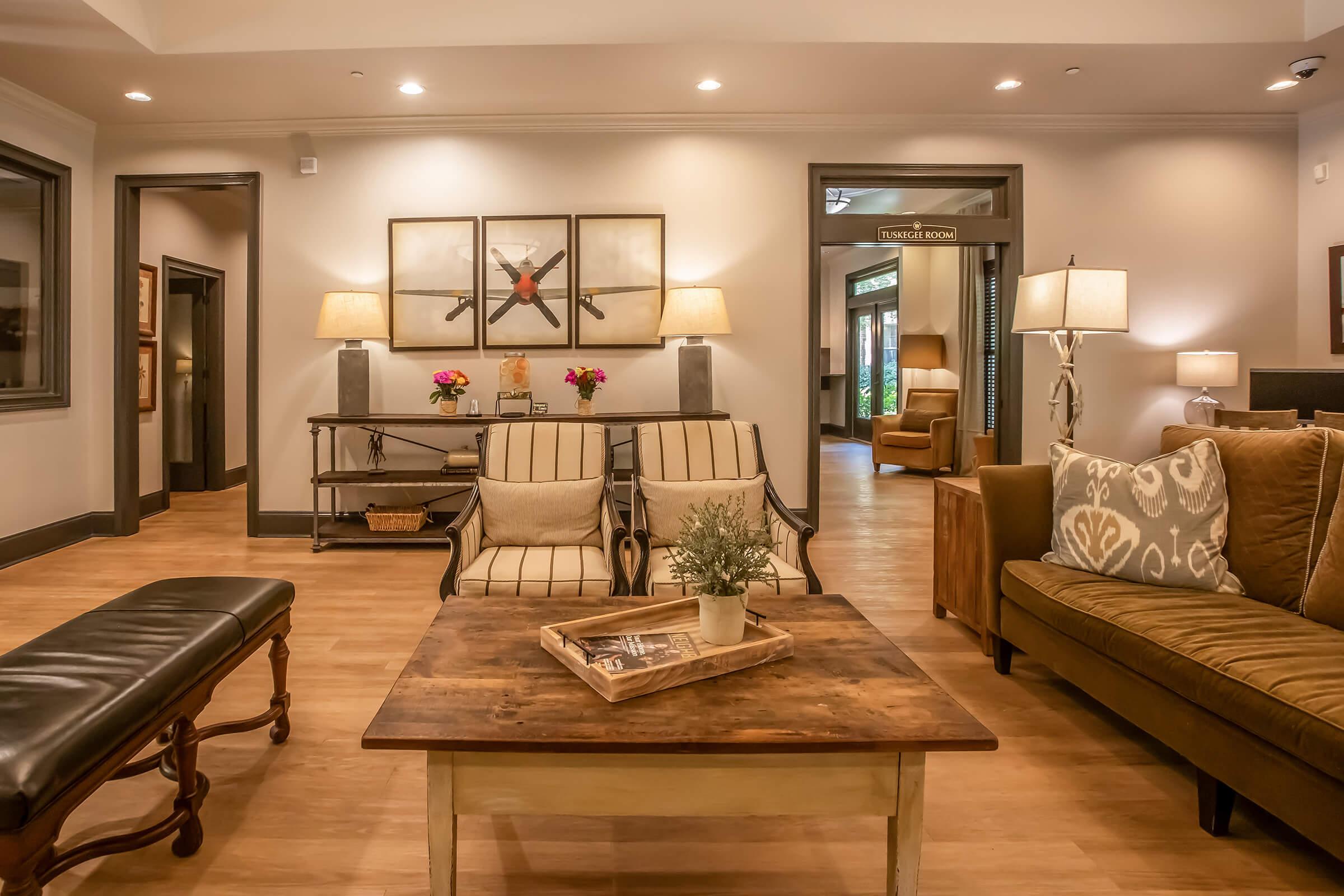 A cozy lounge area featuring two armchairs with striped cushions, a brown sofa with decorative pillows, and a wooden coffee table with a small plant. The space is illuminated by table lamps and has framed artwork on the wall. A doorway leads to a neighboring room labeled "Tuning Room."