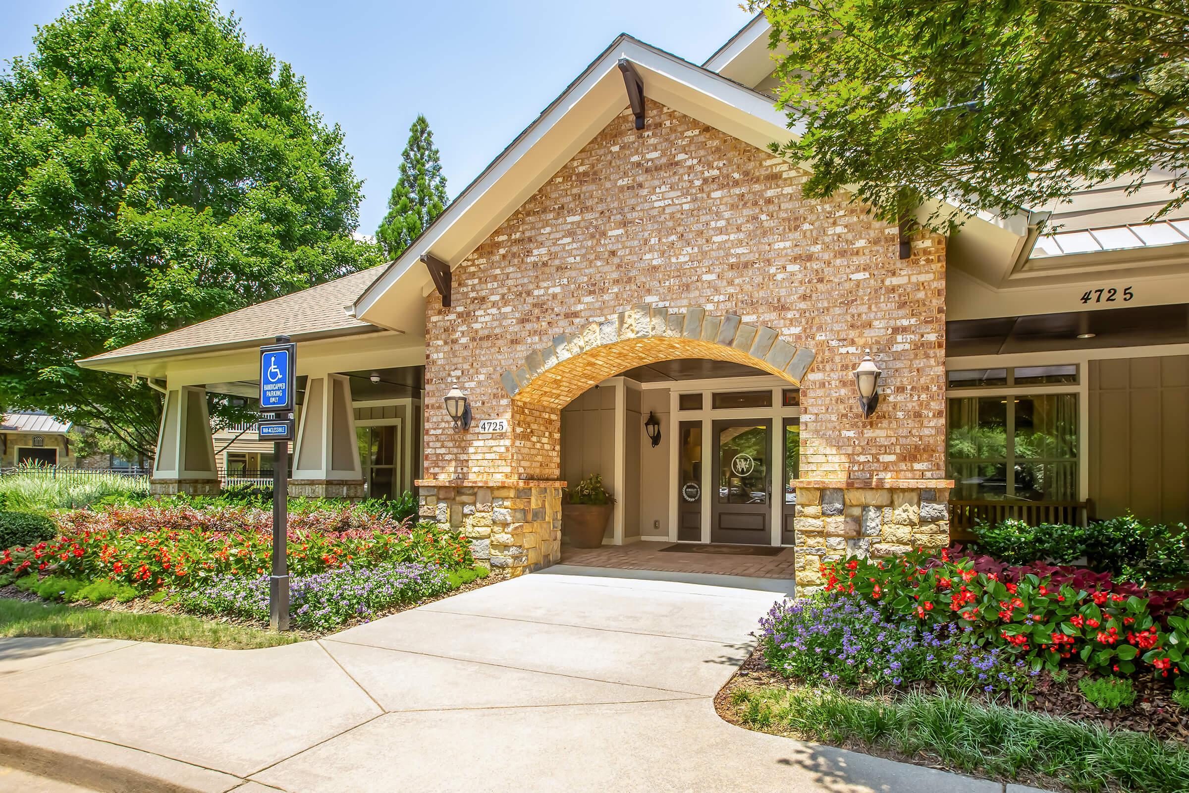 Exterior view of a welcoming building featuring a brick facade, a prominent entryway with an arched design, and landscaped gardens filled with colorful flowers. The pathway leads to the entrance, and there is a designated handicapped parking sign visible nearby, enhancing accessibility.