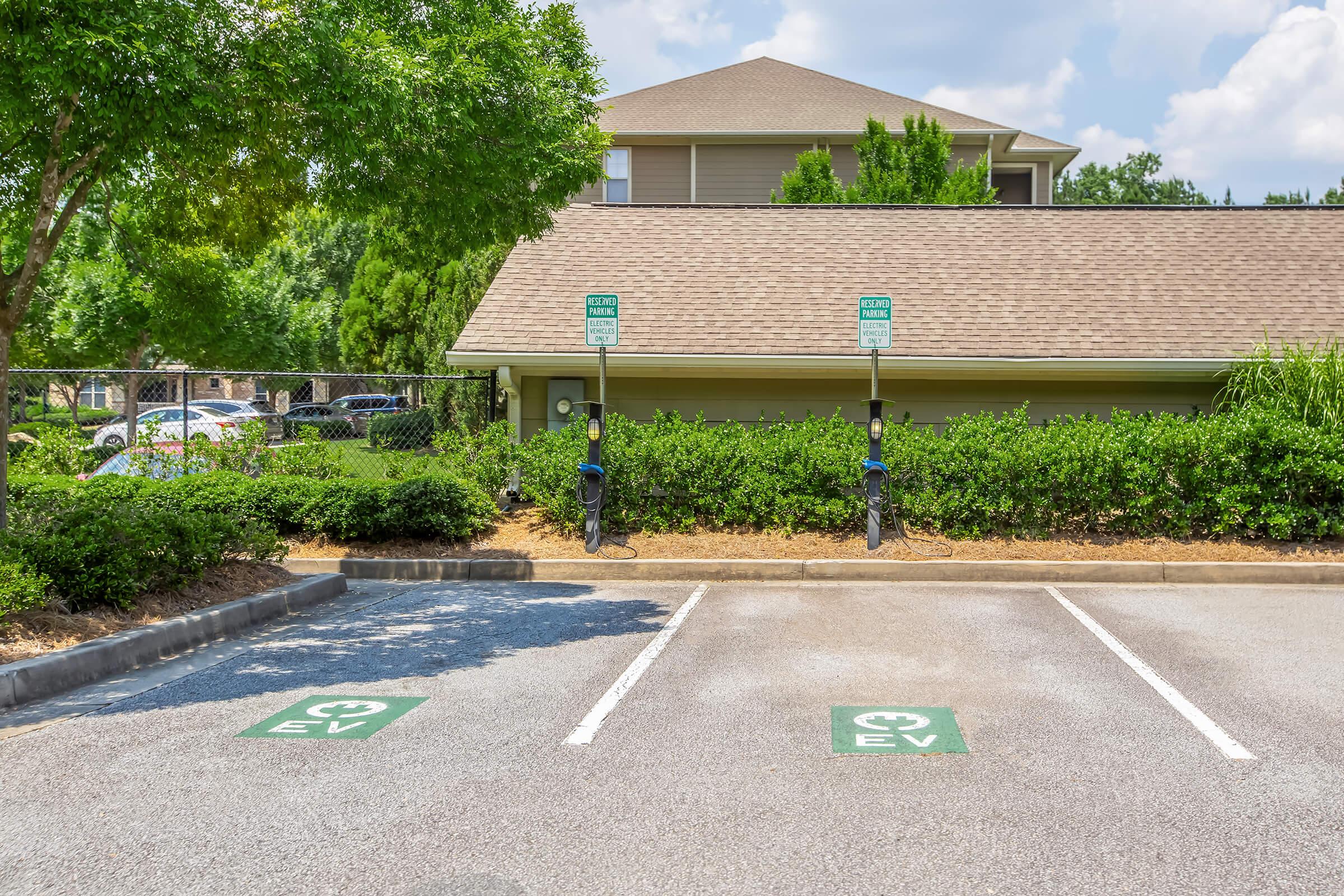 Two electric vehicle (EV) charging stations are positioned in a parking lot, marked with green EV symbols. The background features a building and lush greenery, with clear blue skies above. The area is well-maintained, highlighting accessibility for electric vehicle owners.