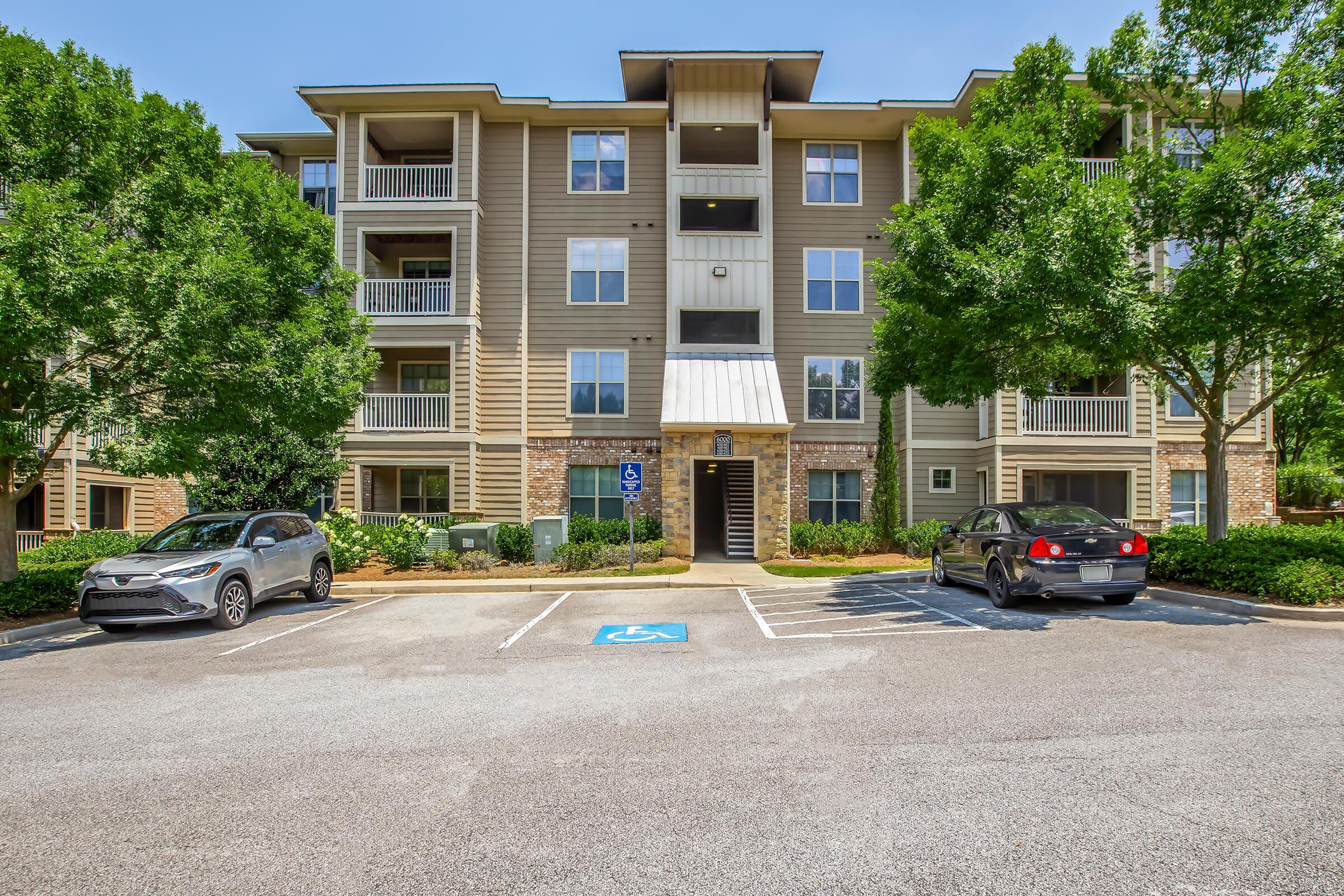 A three-story apartment building with beige siding and balconies. The entrance is at the center, flanked by green shrubs and trees. There are two parked cars in front, one in a disabled parking space. The sky is clear and blue, indicating a sunny day.