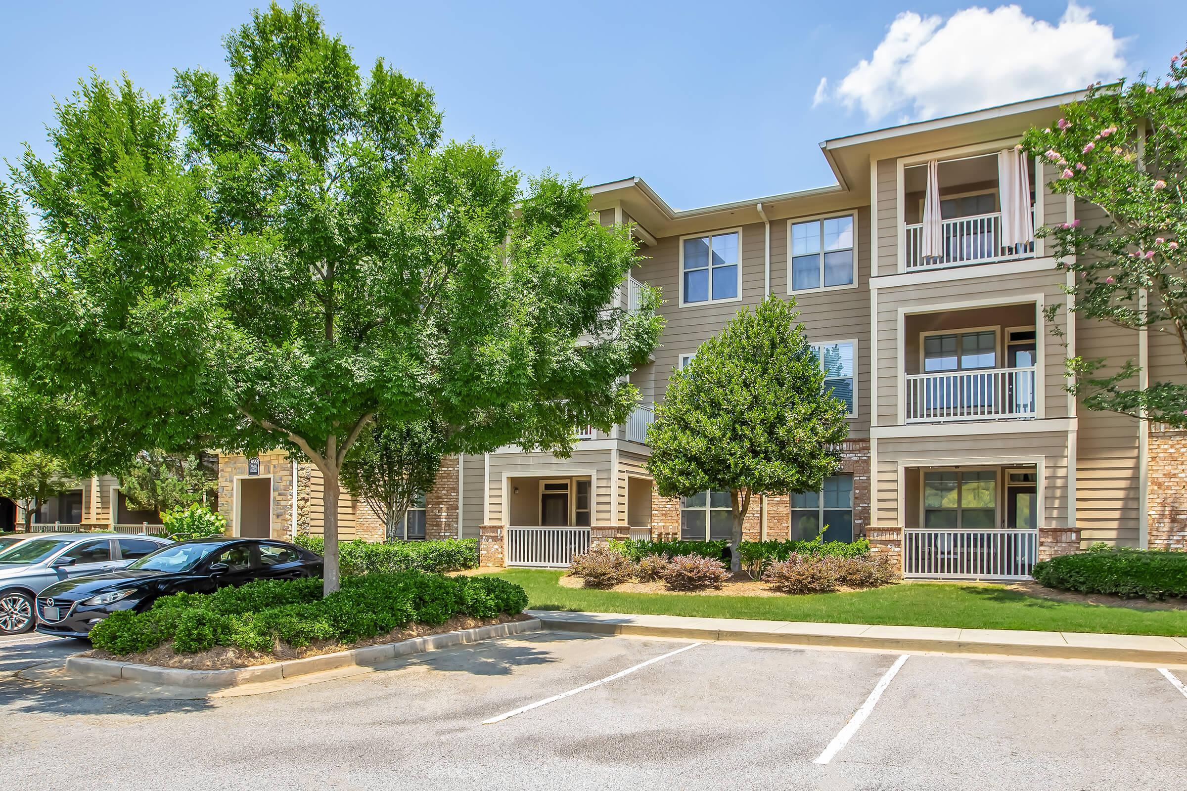Three-story apartment building with stone and beige siding. Balconies on the upper floors, surrounded by well-maintained landscaping, including trees and shrubs. A parking lot is visible in the foreground with several parked cars. The sky is clear with a few clouds.