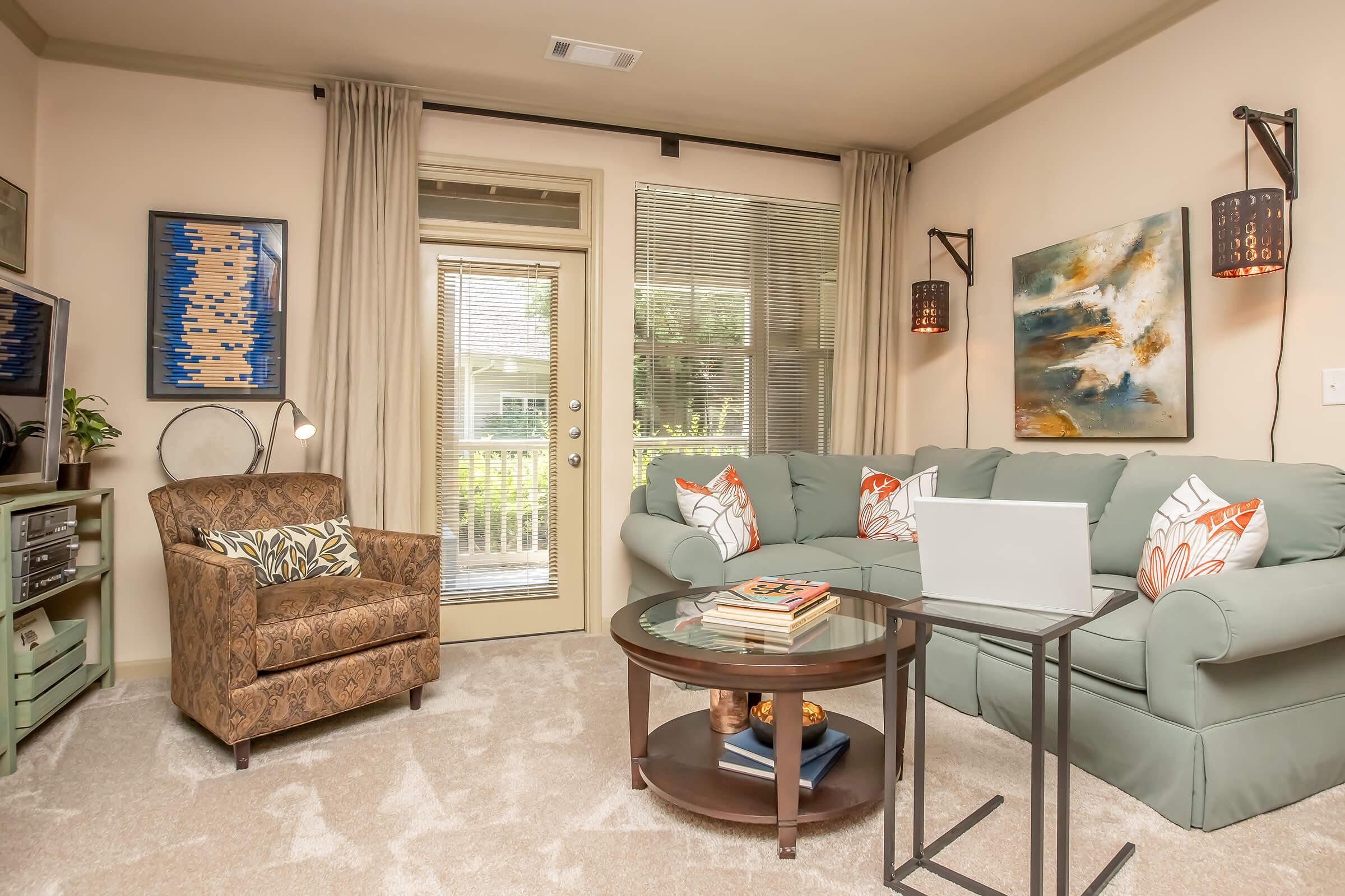 Cozy living room featuring a light green sofa adorned with decorative pillows, a patterned armchair, a round coffee table with books and a decorative bowl, and a TV console. Large windows let in natural light, complemented by sheer curtains and wall art. A door leads to an outdoor area.