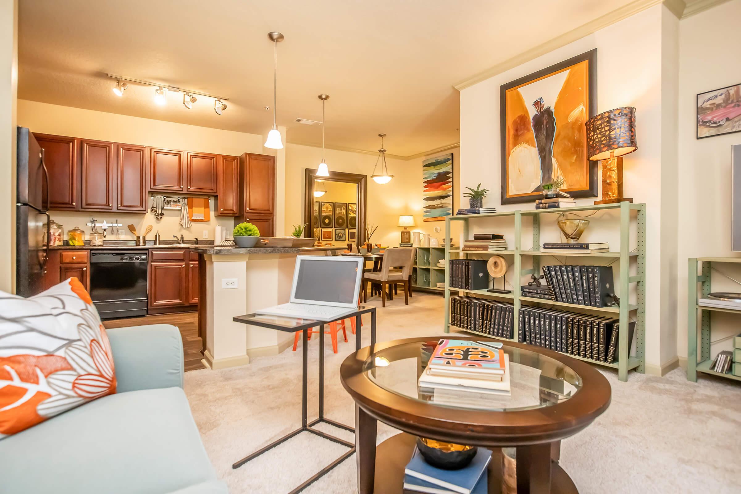 A cozy living space featuring a light-colored sofa with orange and blue cushions, a small coffee table with magazines, and a laptop. In the background, a modern kitchen with wooden cabinets, a dining area with a table and chairs, and decorative shelves filled with books and plants. Warm lighting enhances the inviting atmosphere.