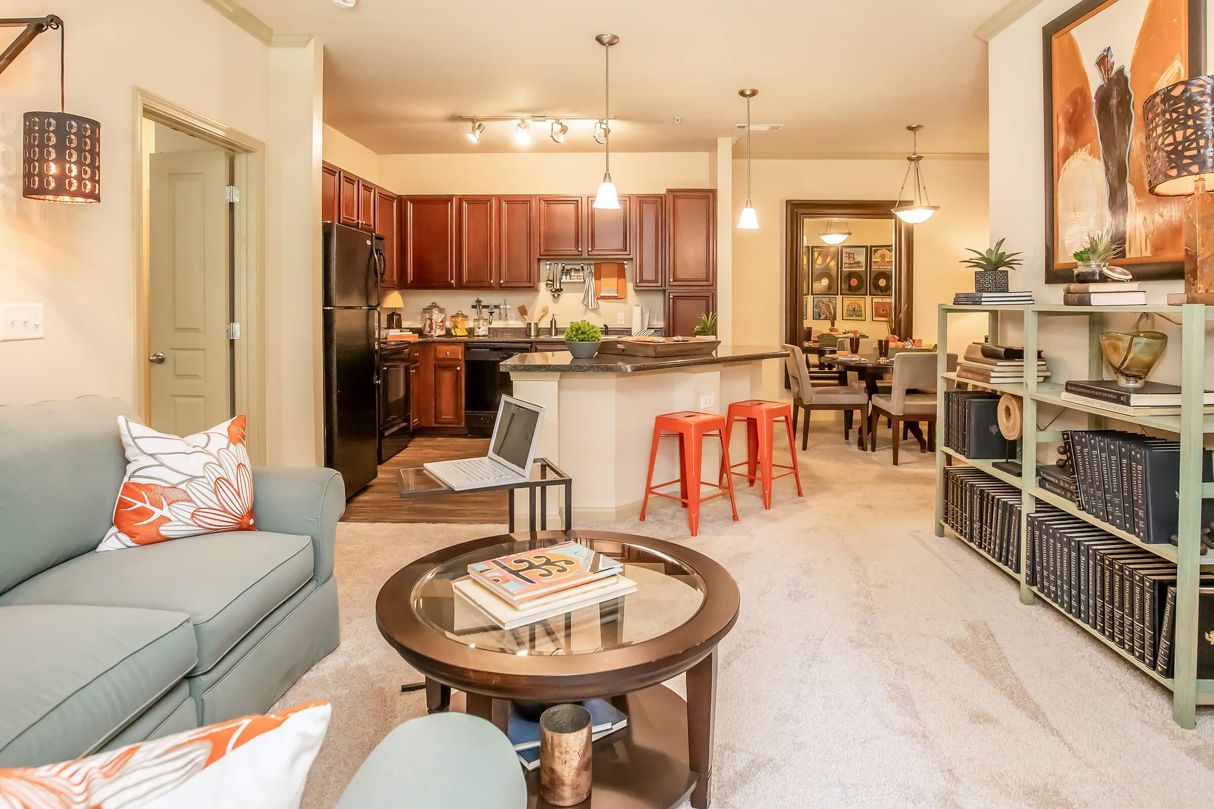 A cozy living area showcasing a light green sofa with orange-patterned cushions, a round coffee table with a stack of magazines, and an open kitchen with dark wood cabinets. The dining area features a small table and red stools, complemented by decorative wall art and plants, creating a warm and inviting atmosphere.