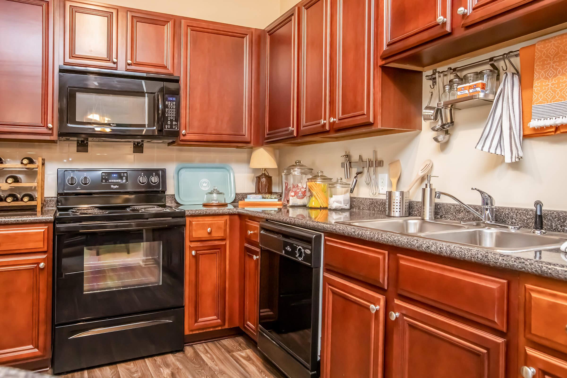 Modern kitchen featuring dark wood cabinetry, granite countertops, and stainless steel appliances. The setup includes a black stove, microwave, and dishwasher, as well as a variety of kitchen utensils and decorative items. A light green kitchen appliance and a fruit jar add a pop of color to the space.