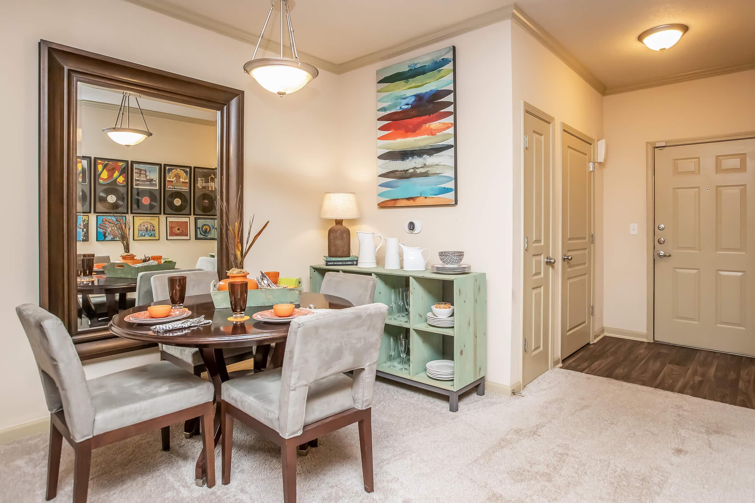 A cozy dining area featuring a round wooden table set with plates and glasses, surrounded by four upholstered chairs. Adjacent is a green sideboard with decor items. A large mirror reflects the space, and colorful artwork hangs on the wall, while a welcoming front door leads outside. The carpeted floor adds warmth.