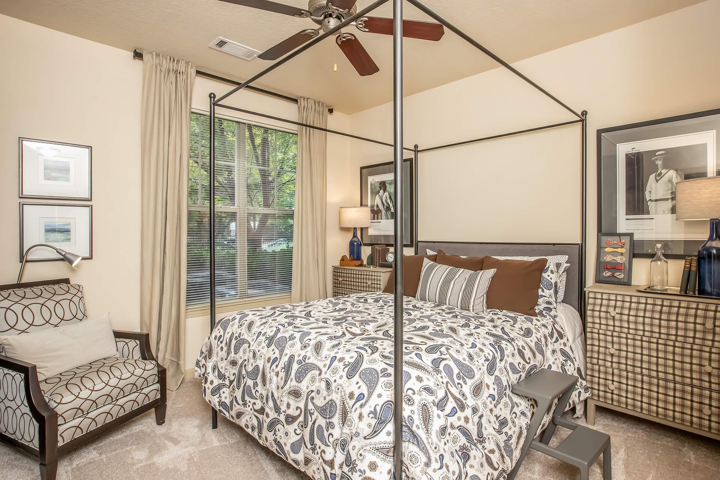 A cozy bedroom featuring a four-poster bed, adorned with patterned bedding. There's an armchair in the corner, framed artwork on the walls, and a bedside table with a lamp. Large windows allow natural light to enter, and neutral tones create a warm, inviting atmosphere.