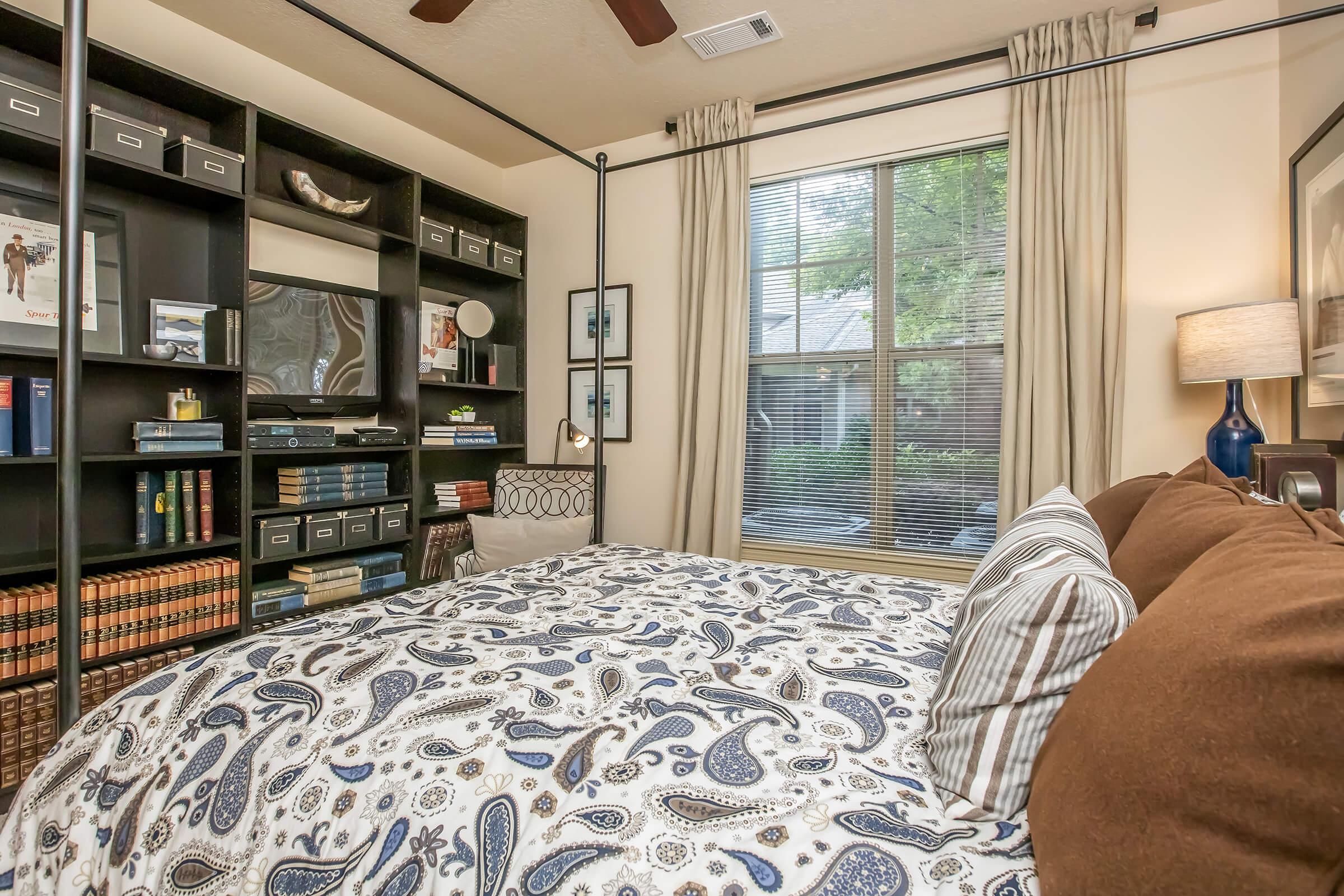 A cozy bedroom featuring a patterned bedspread, decorative pillows, and a dark wood bookshelf filled with books and storage boxes. A lamp sits on a bedside table, and large windows with curtains allow natural light to illuminate the space, creating a warm and inviting atmosphere.