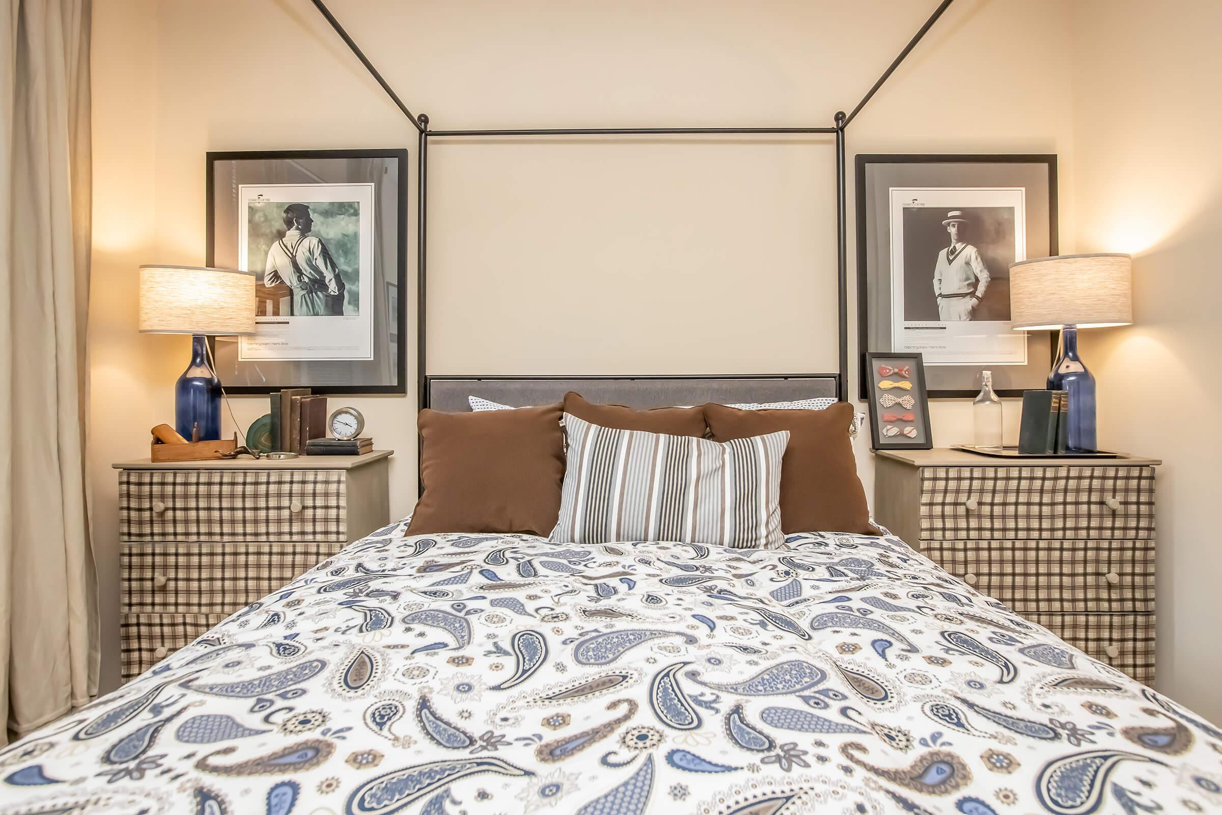 A cozy bedroom featuring a styled bed with a patterned comforter. Two bedside tables with decorative items and lamps flank the bed. Above the headboard are framed black-and-white photographs. The walls are a soft, neutral color, creating a warm and inviting atmosphere.