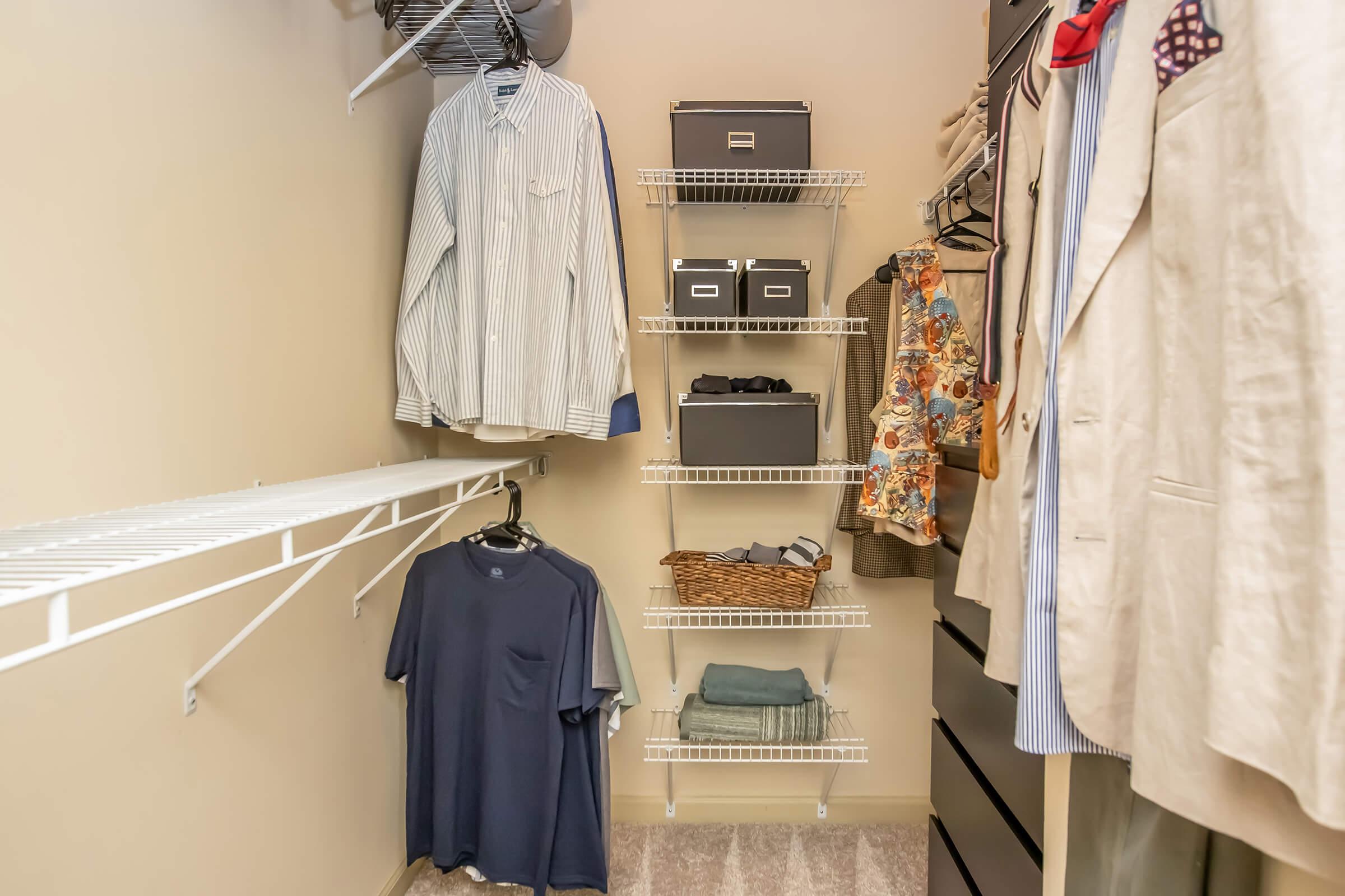 A well-organized closet featuring hanging shirts, a collection of neatly folded items, storage boxes, and a decorative basket on shelves. The space is designed for efficient use, with light-colored walls and clean, minimal decor.