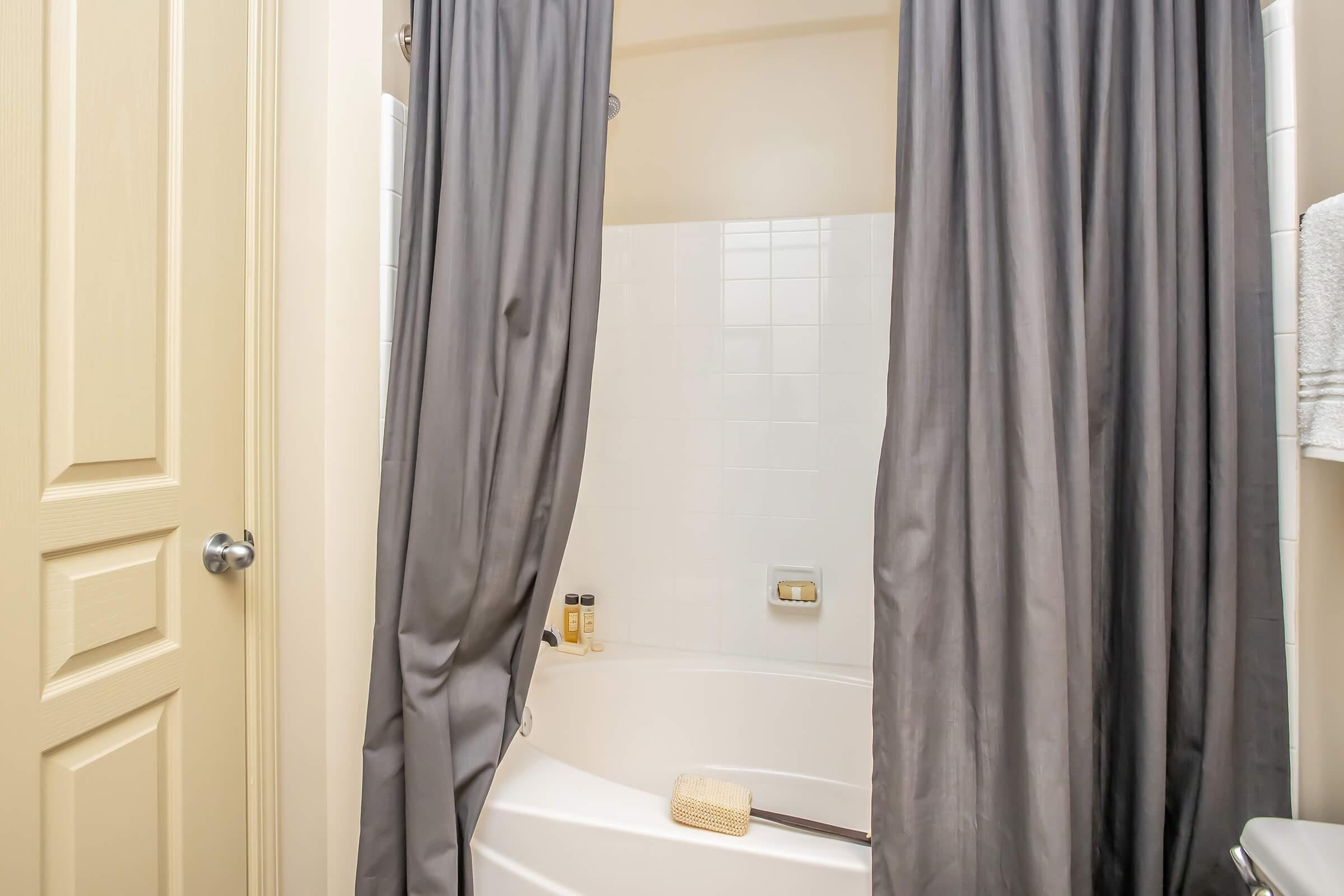 A bathroom with a shower area partially hidden by gray curtains. The white tiled wall features a small shelf with toiletries. The tub is clean and has a wooden bath brush resting on its edge. A closed door with a simple design is visible on the left side.