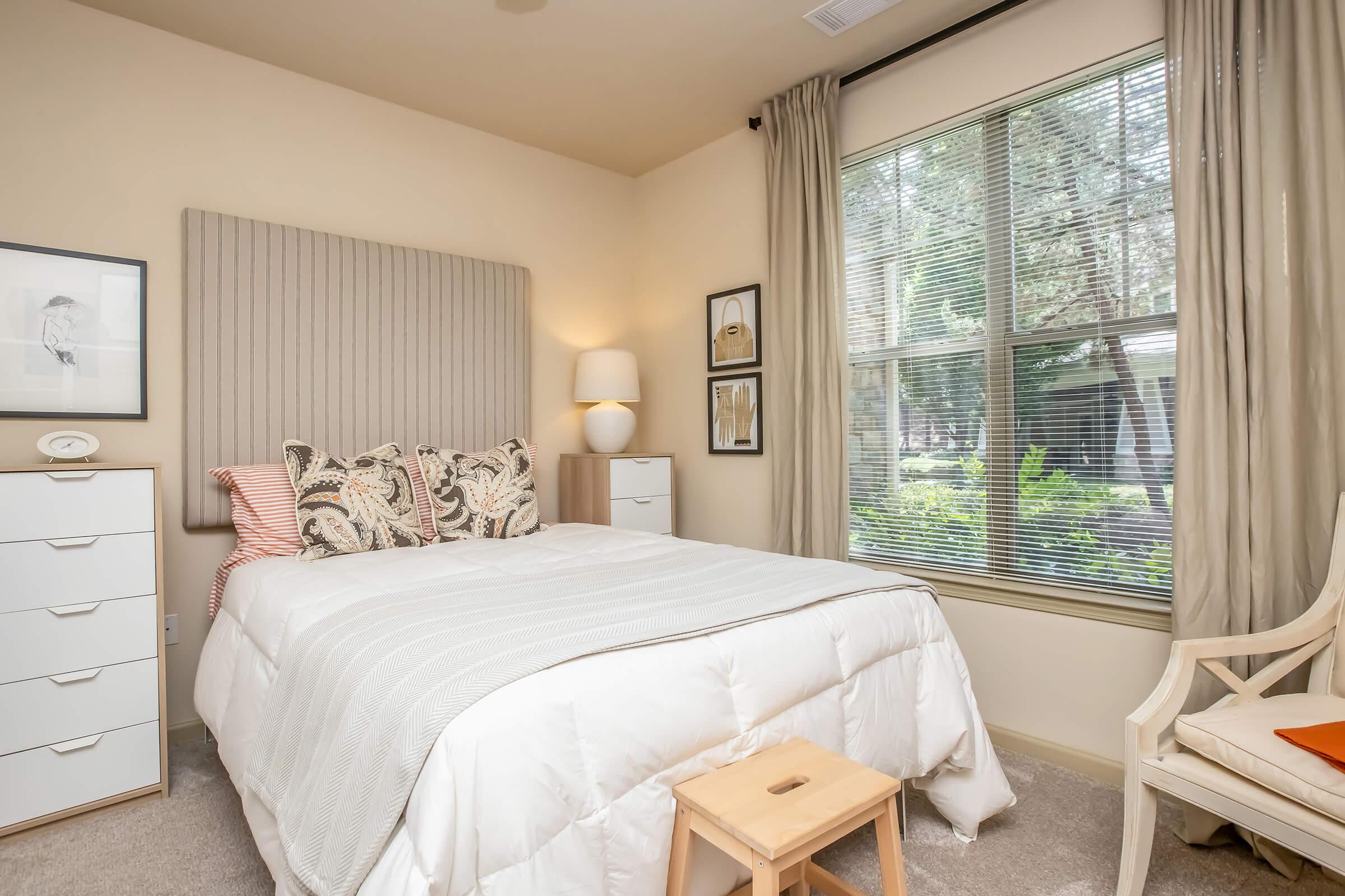 Cozy bedroom featuring a queen-sized bed with decorative pillows, a light-colored headboard, and a white comforter. A side table with a lamp sits next to the bed. Large window with sheer curtains lets in natural light, and a white chest of drawers is positioned nearby, adding to the room's inviting atmosphere.