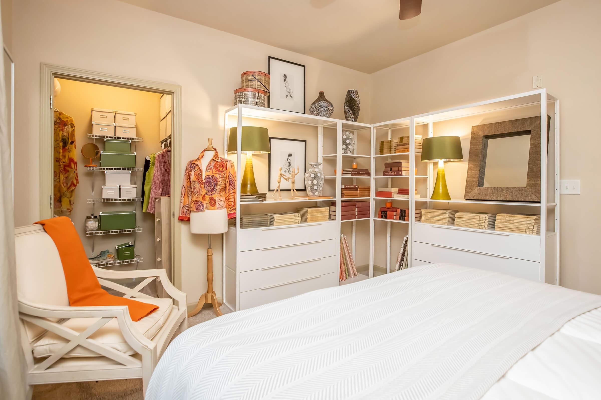 A cozy bedroom featuring a white bed with a textured blanket, a stylish wardrobe with drawers and lamps, and decorative items on display. A small closet with organized boxes is visible in the background, along with a clothing rack and a mannequin. The room has warm, inviting colors and a light ambiance.