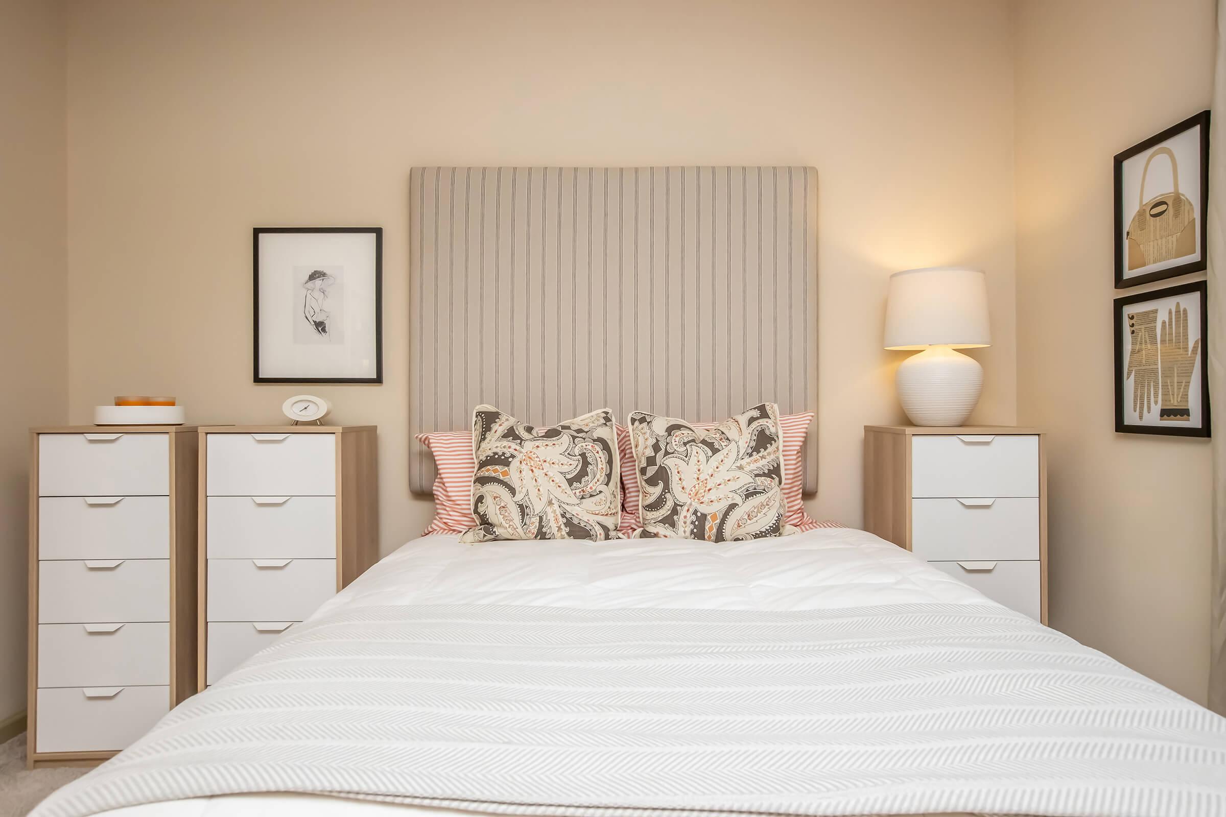 A cozy bedroom featuring a neatly made bed with patterned pillows, a light-colored headboard, and white bedding. Flanking the bed are two modern dressers with multiple drawers. On the walls are framed artworks, and a bedside lamp casts a warm glow, creating a welcoming atmosphere.
