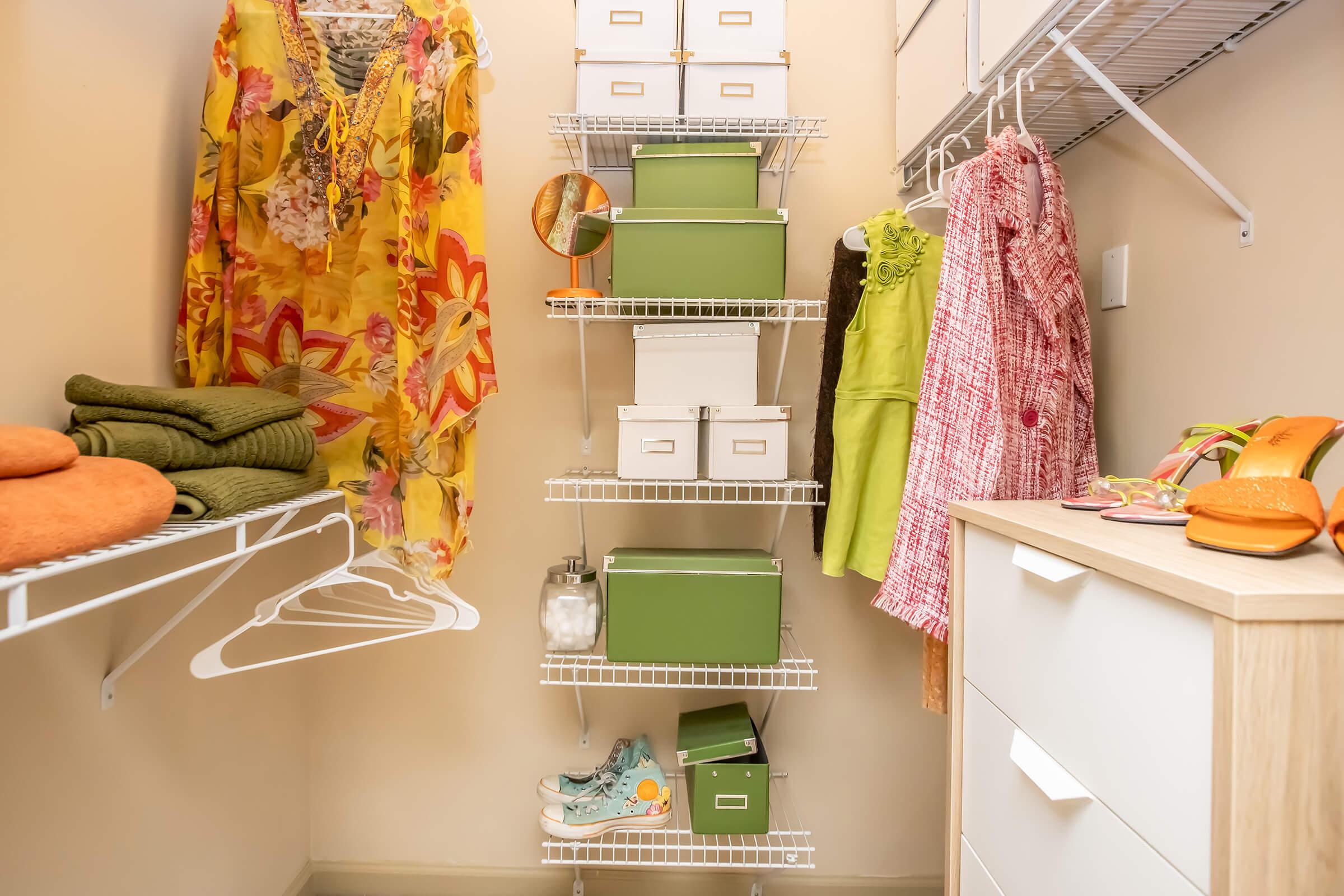 A well-organized closet featuring colorful clothing items like a floral dress and a lime green outfit. Shelves hold storage boxes in green and white, along with a pair of casual shoes and accessories like a bright bag. Freshly folded towels are neatly stacked on the lower shelf.
