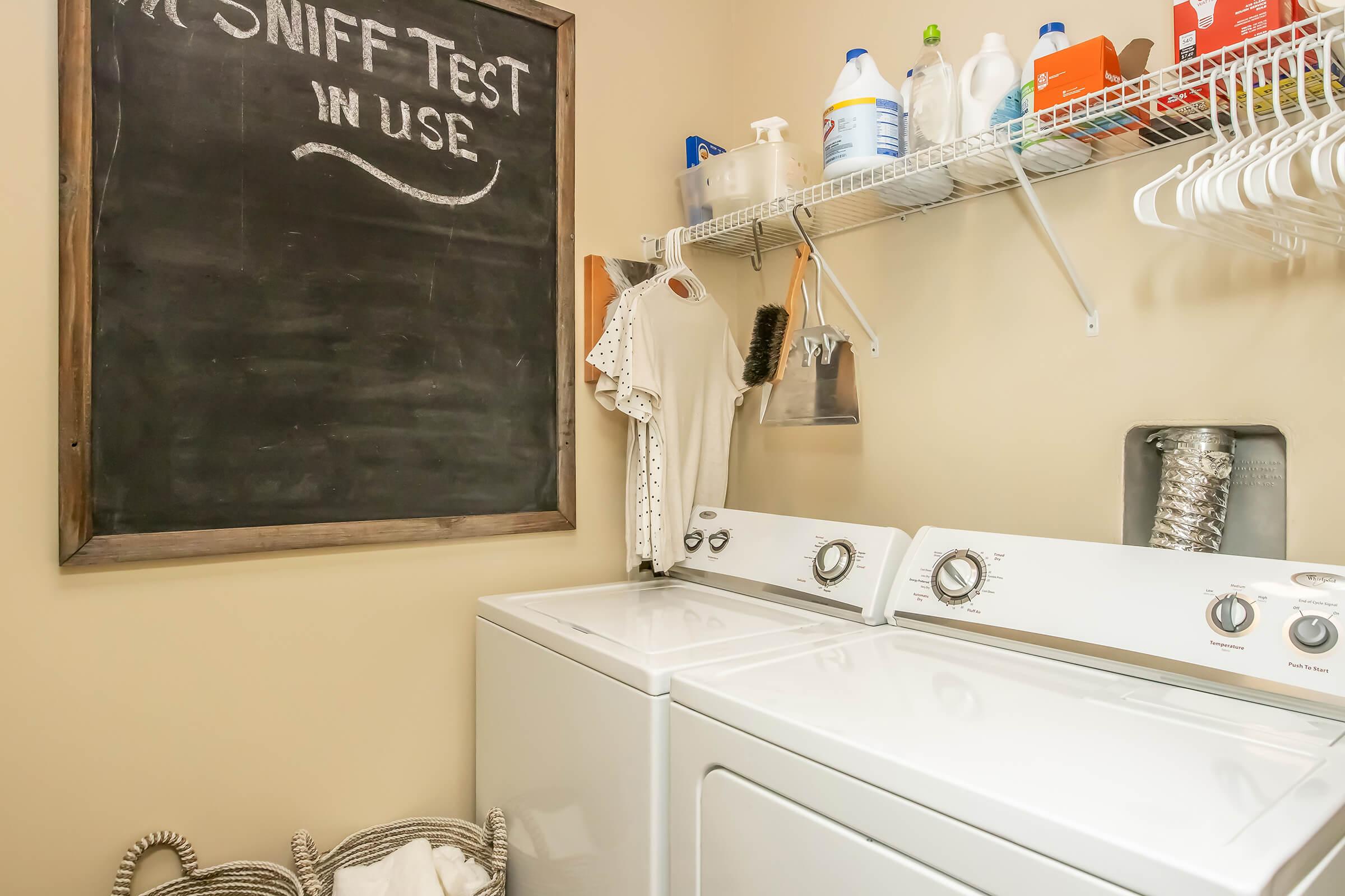A laundry room featuring a black chalkboard with "SNIFF Test IN USE" written on it, a washing machine and dryer, cleaning supplies on shelves, and clothes hanging on hooks. A wicker basket is also present, adding to the organized and clean appearance of the space.