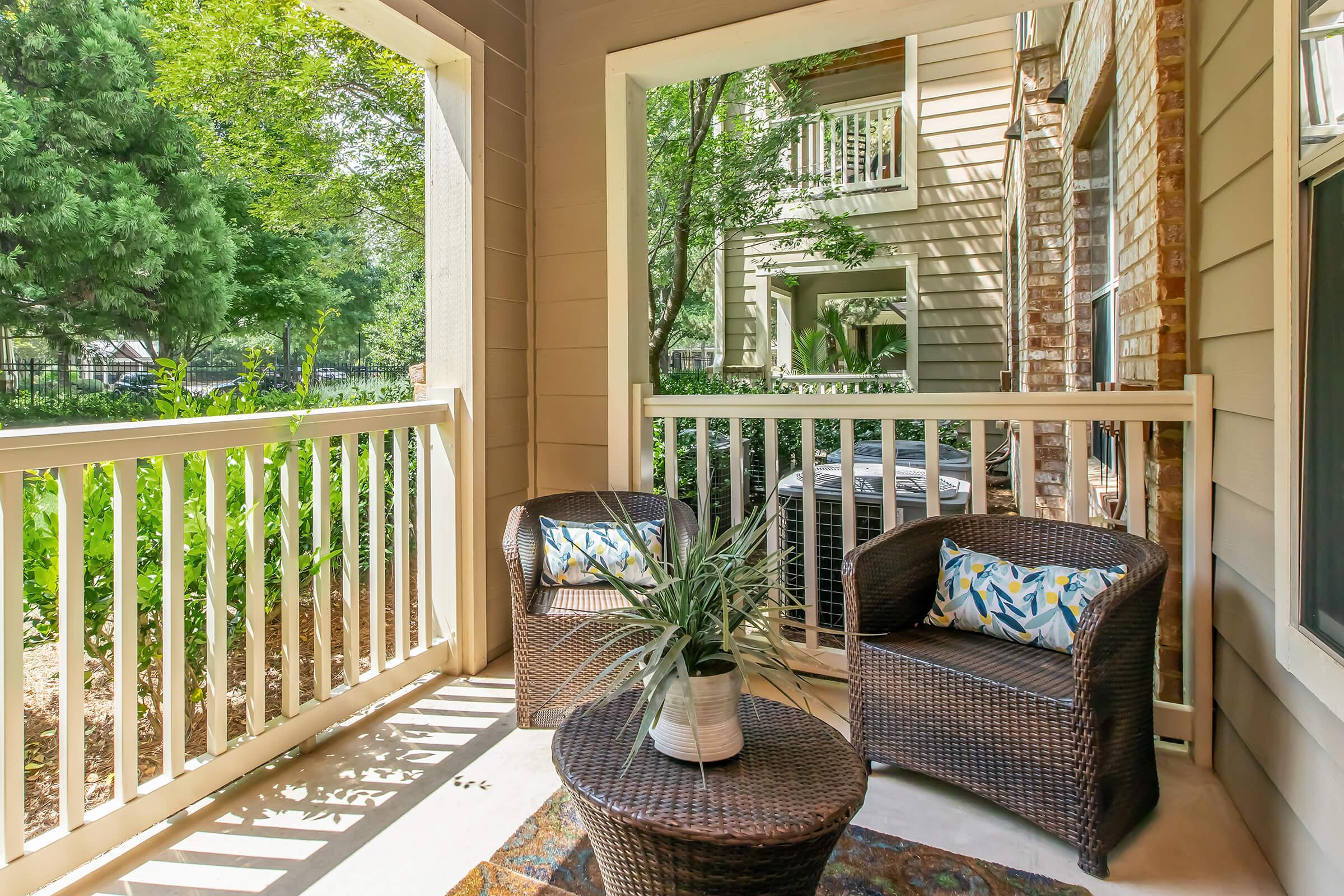 A cozy porch area featuring two wicker chairs with decorative pillows and a small round table. A potted plant is on the table, and there is a view of greenery outside along with nearby building architecture. The space is bright and inviting, perfect for relaxation.