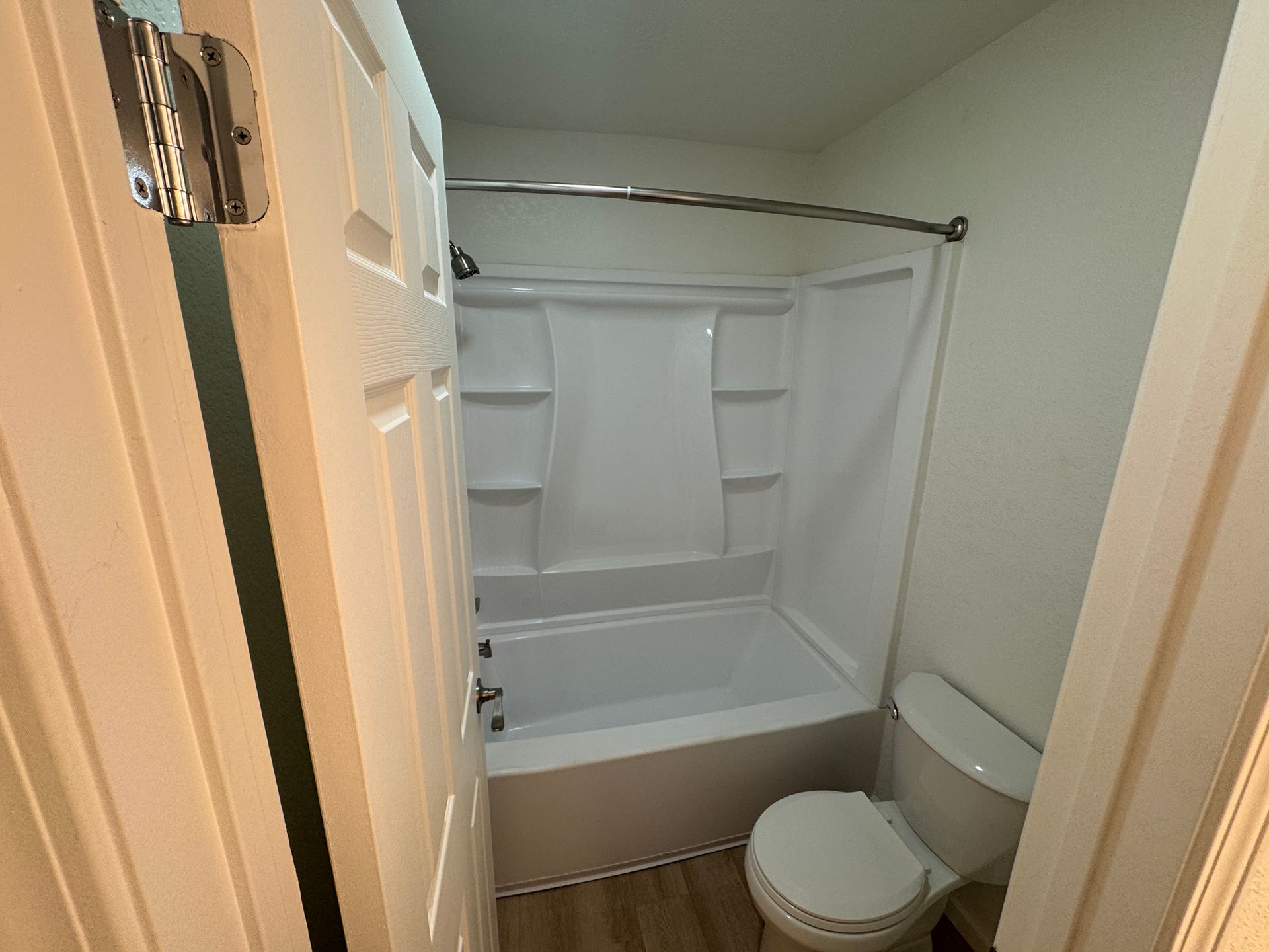 A small bathroom featuring a shower-tub combination, a toilet, and light-colored walls. The door is closed, and the flooring appears to be light wood. The overall space is simple and functional.