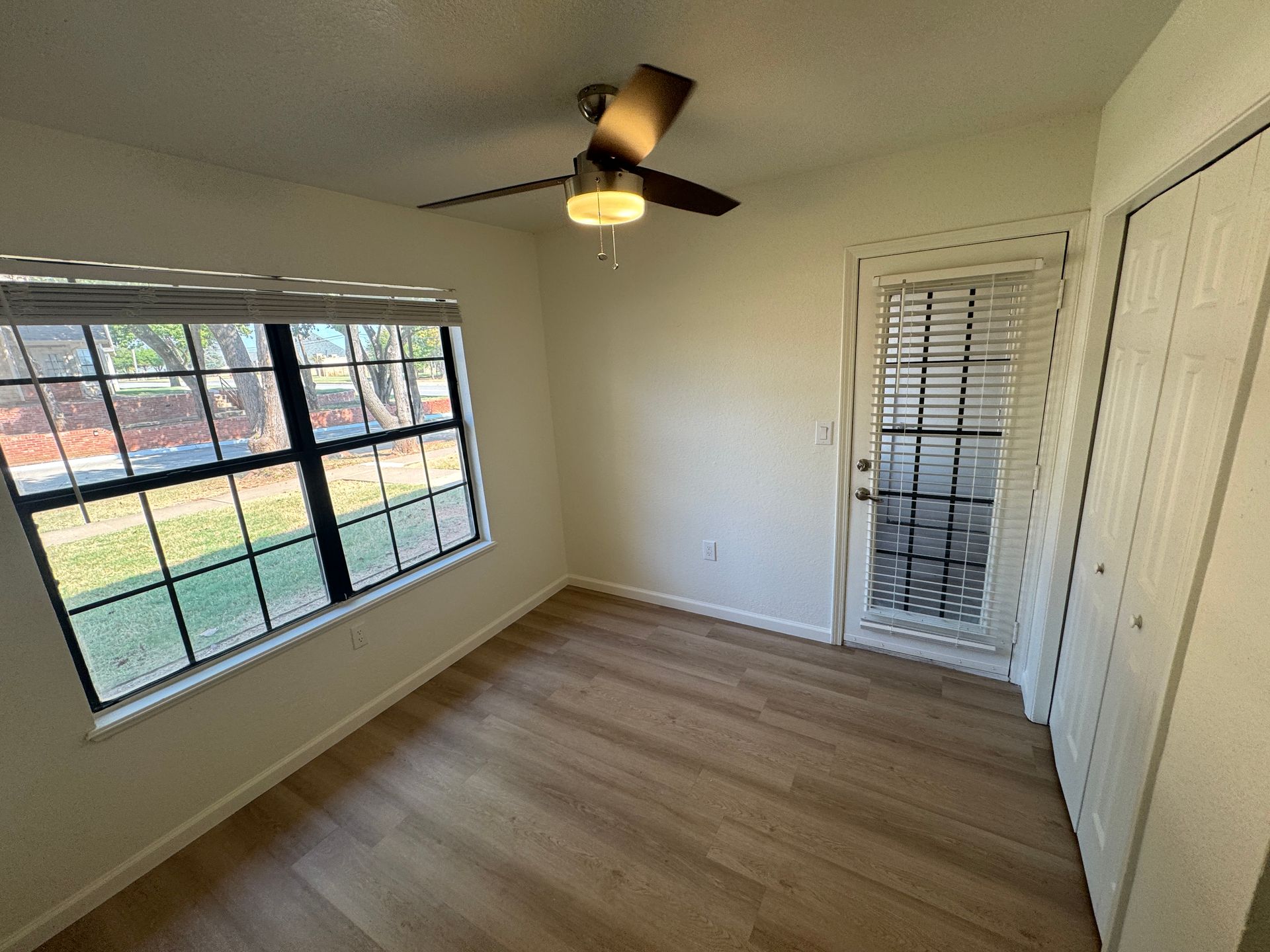 A bright, empty room featuring a ceiling fan and large windows with black frames. The flooring is light wood, and there is a door leading outside on one side. The walls are painted white, creating a clean and airy atmosphere. Sunshine illuminates the space through the windows.