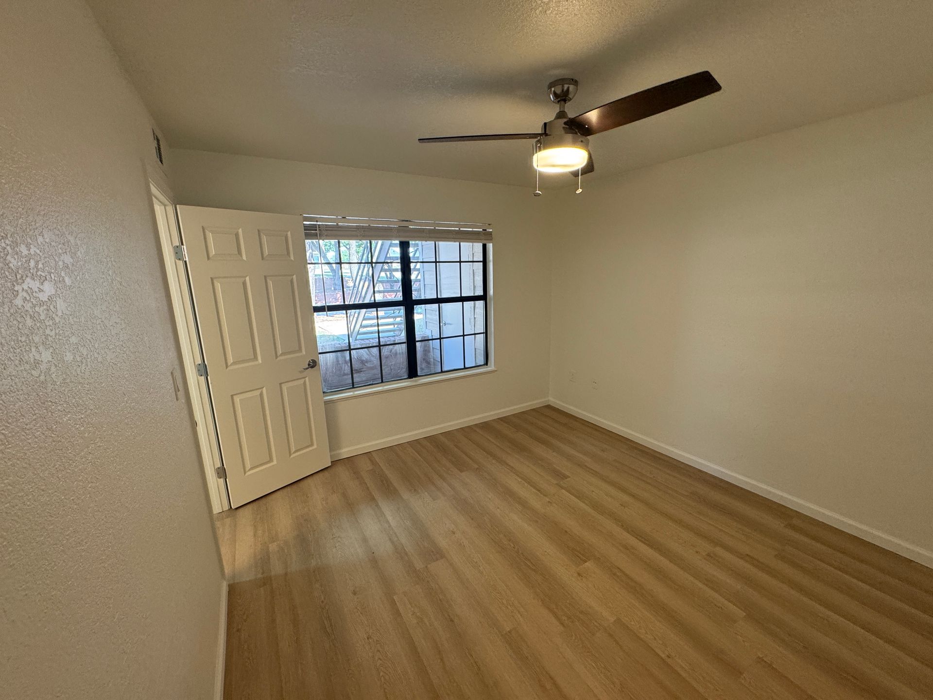 Empty room with light-colored walls and a window allowing natural light. There’s a ceiling fan with a light fixture, and the floor is made of light wood. An open door leads to another space, enhancing the room's spacious feel.