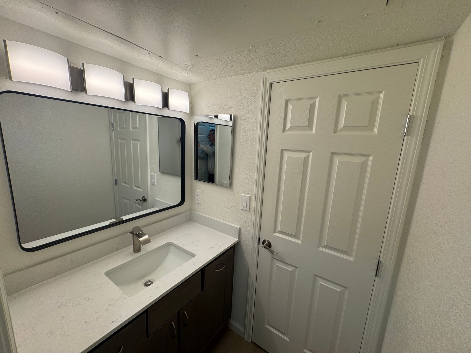 A well-lit bathroom with a modern design. It features a rectangular mirror above a white sink with a single faucet. The light fixtures above the mirror are sleek and contemporary. To the right, there is a door leading to another room, and the walls are painted a light color, enhancing the space's brightness.