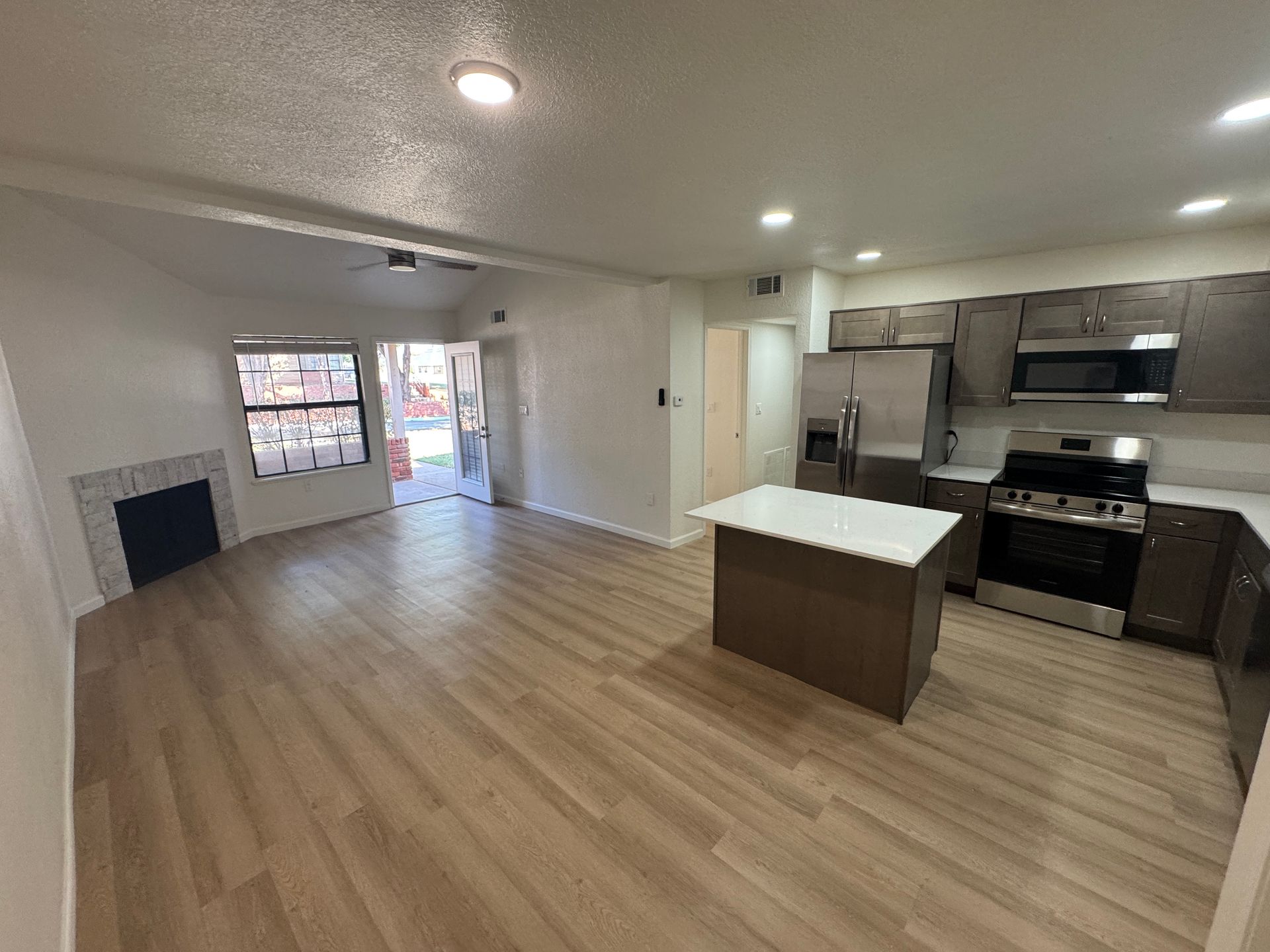 A modern open-concept kitchen and living area featuring light wooden flooring, a kitchen island with a white countertop, stainless steel appliances, and dark wood cabinetry. A large window provides natural light, and a doorway leads to an outdoor area. A fireplace is visible in the corner of the living space.