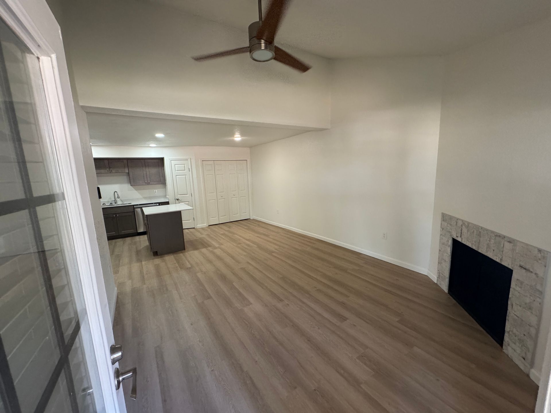 A spacious, empty living area featuring light wood flooring, a ceiling fan, and a stone fireplace. Adjacent to the living space is a modern kitchen with dark cabinetry. The room has neutral walls and ample natural light, creating a bright and open atmosphere.