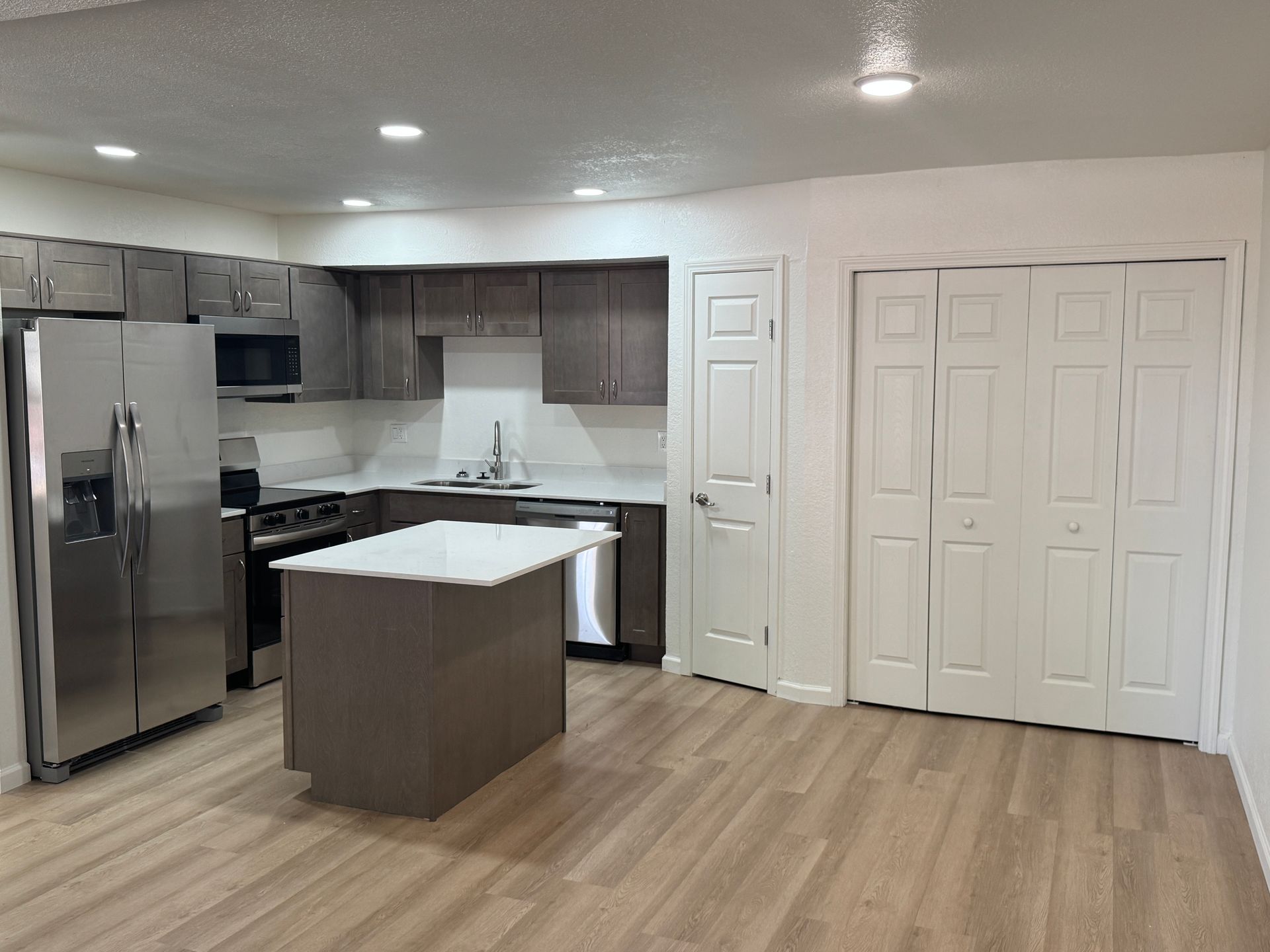 Modern kitchen featuring stainless steel appliances, dark wooden cabinets, and a large island with a white countertop. The open layout includes a pantry closet and neutral-colored walls, complemented by warm lighting. Vinyl flooring adds a contemporary touch to the space.
