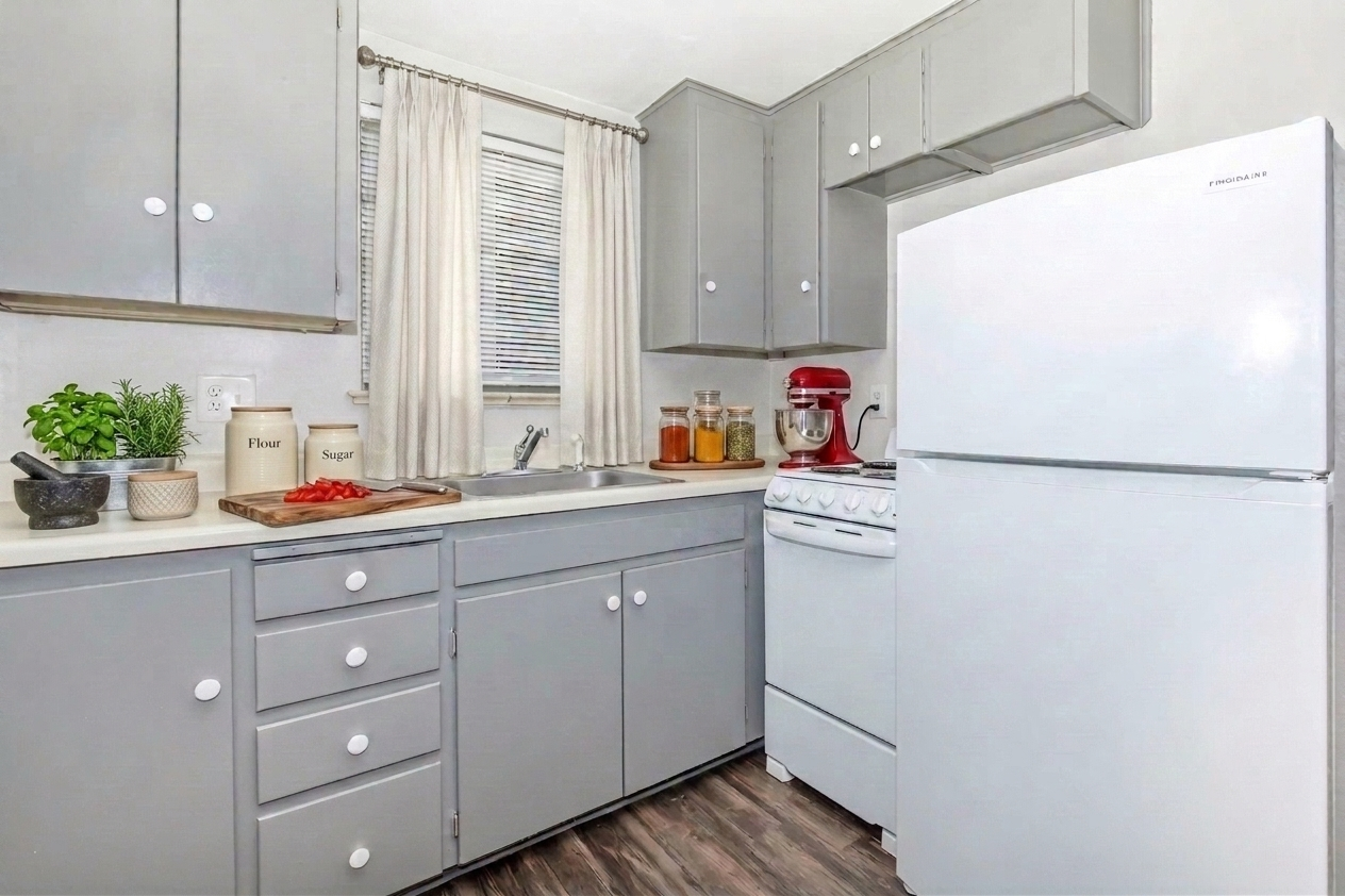 A modern kitchen featuring gray cabinets and drawers, a white refrigerator, a sink with a window above it, and a red stand mixer. Countertop items include a small plant, jars, and a cutting board. The flooring is wood-like, enhancing the cozy atmosphere. Light curtains drape over the window.