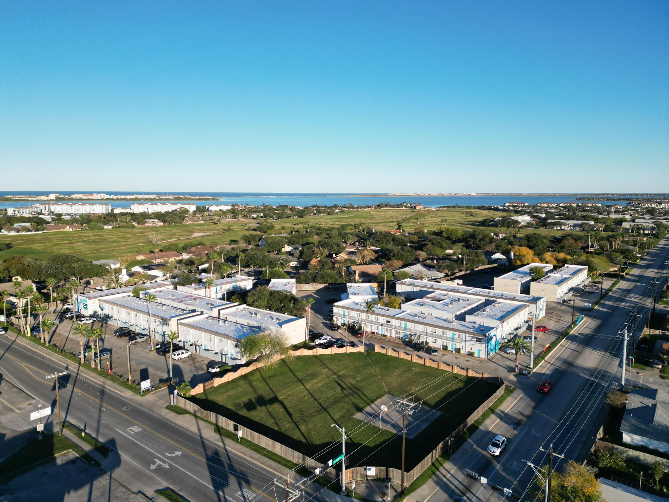 Aerial view of a coastal area featuring a cluster of low-rise buildings, a green lawn, and a nearby road. In the background, there is a body of water with distant land and buildings under a clear blue sky. The scene showcases a blend of urban and natural elements.