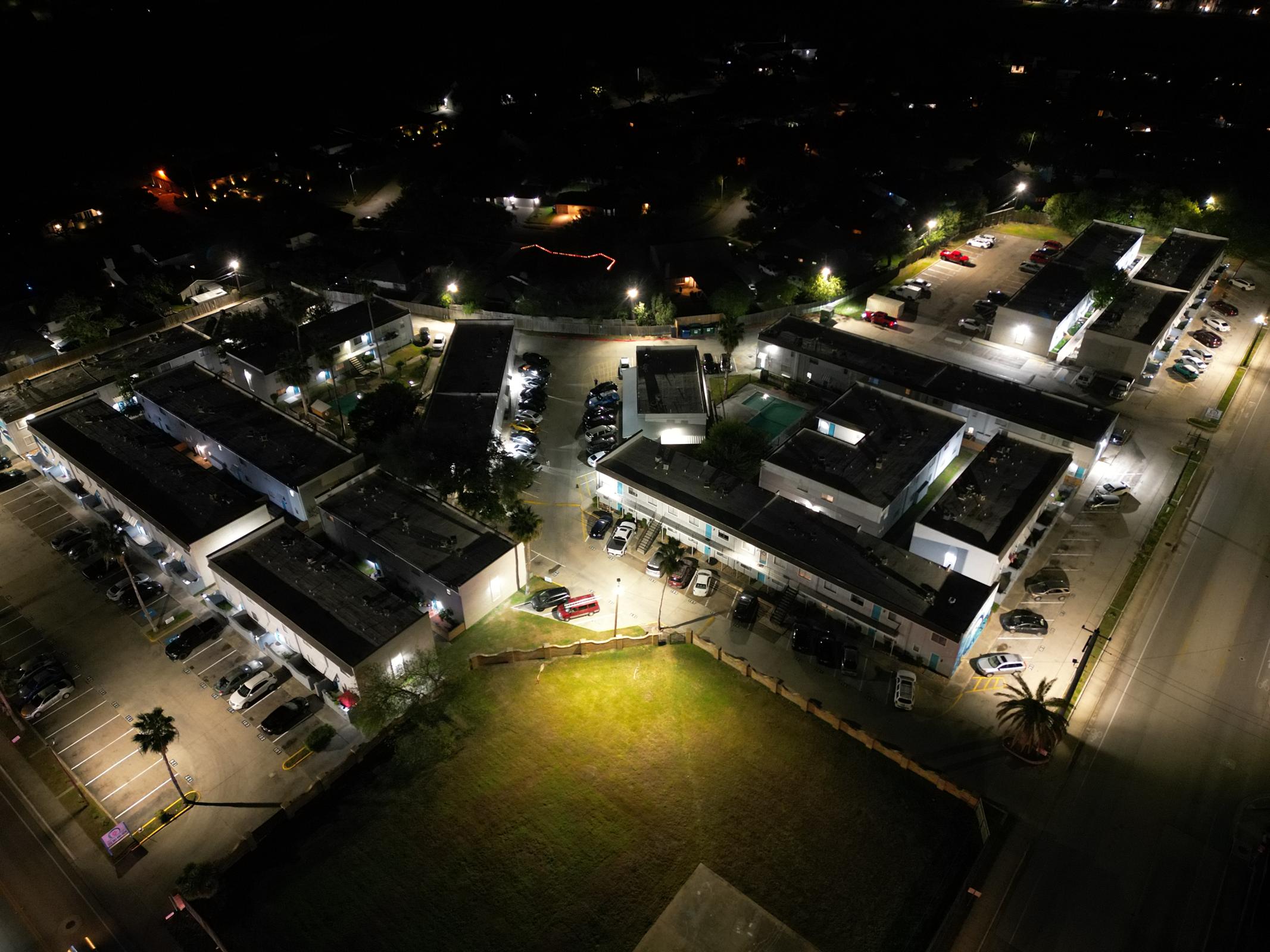 Aerial view of a commercial complex at night, featuring several low-rise buildings with illuminated rooftops and surrounding parking lots. Cars are parked in organized rows, while nearby residential areas are dimly lit. A green field is visible to the left, contrasting with the bright lights of the complex.