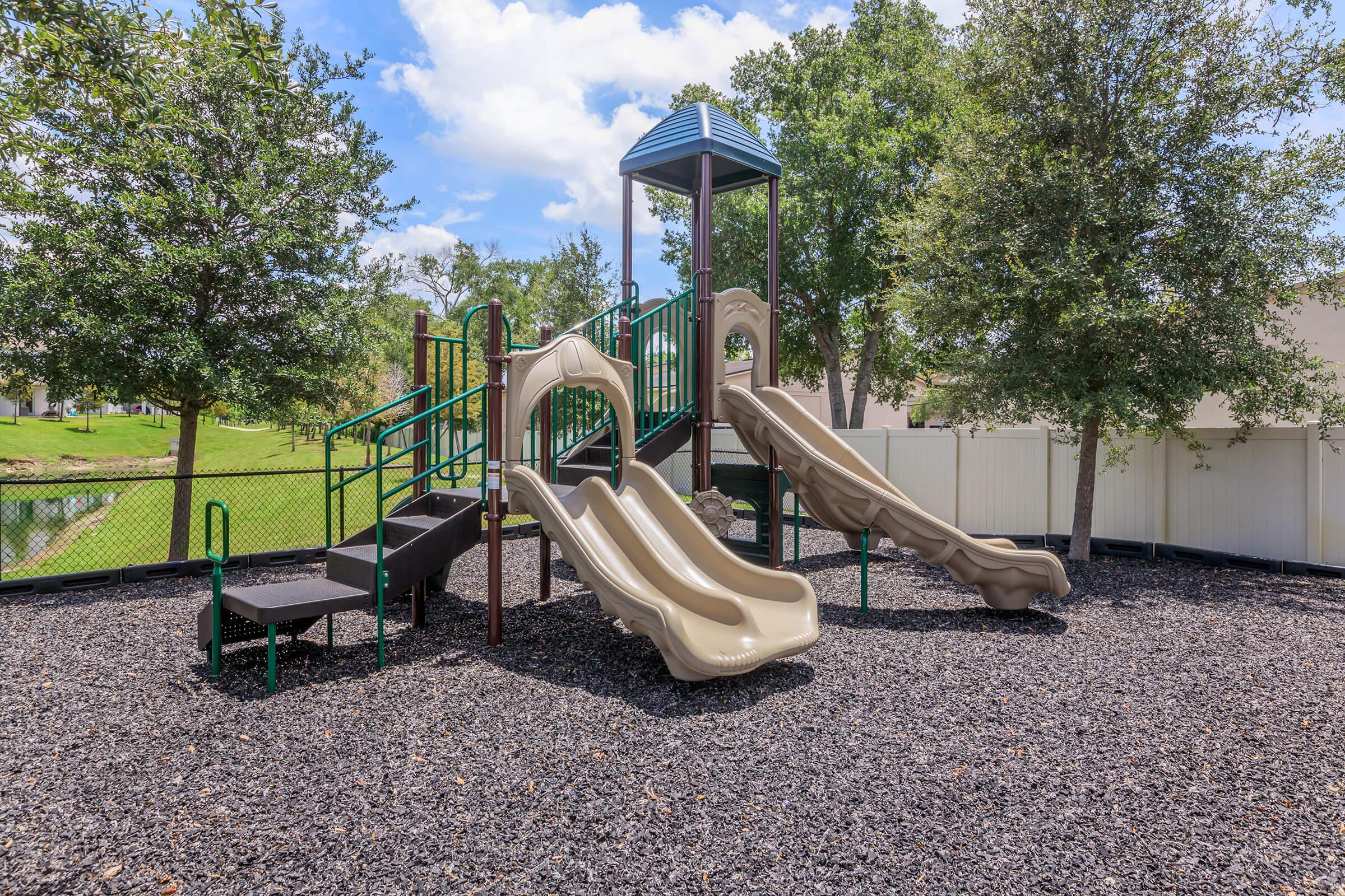 A playground featuring two tan slides, climbing structures, and a safety railing, situated on a bed of black mulch. Surrounding greenery includes trees and a fence, with a serene backdrop of a pond and blue sky with fluffy clouds above.