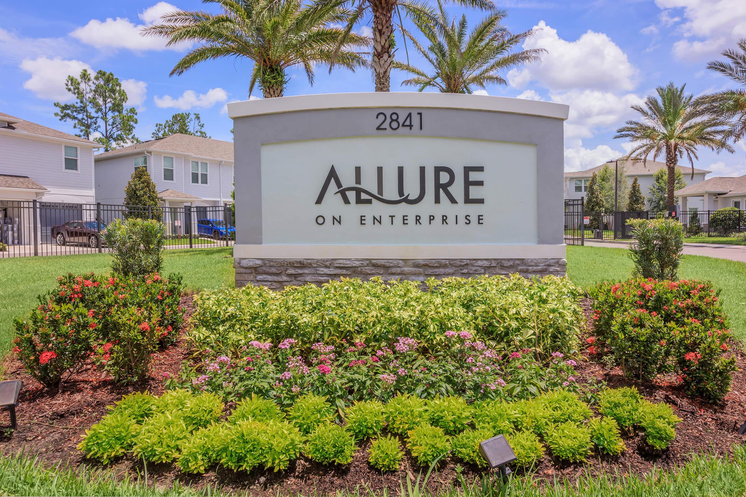 Sign for "Allure on Enterprise," located at 2841, featuring a modern design with landscaped garden including colorful flowers and greenery. The background includes palm trees and blue skies, creating a welcoming atmosphere.