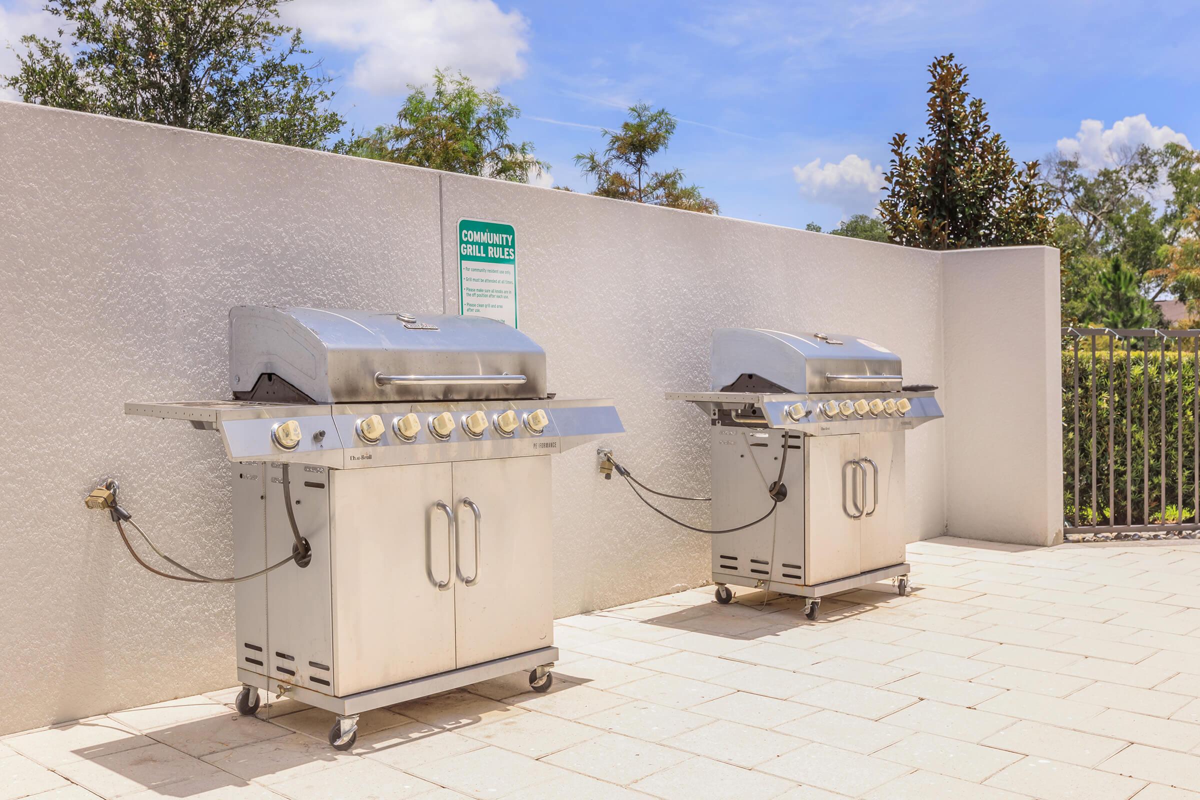 Two stainless steel grills placed side by side in a community outdoor area. They are backed by a light-colored wall, with a sign regarding grill rules visible above. The setting features blue skies and greenery in the background, with a tiled floor below the grills.