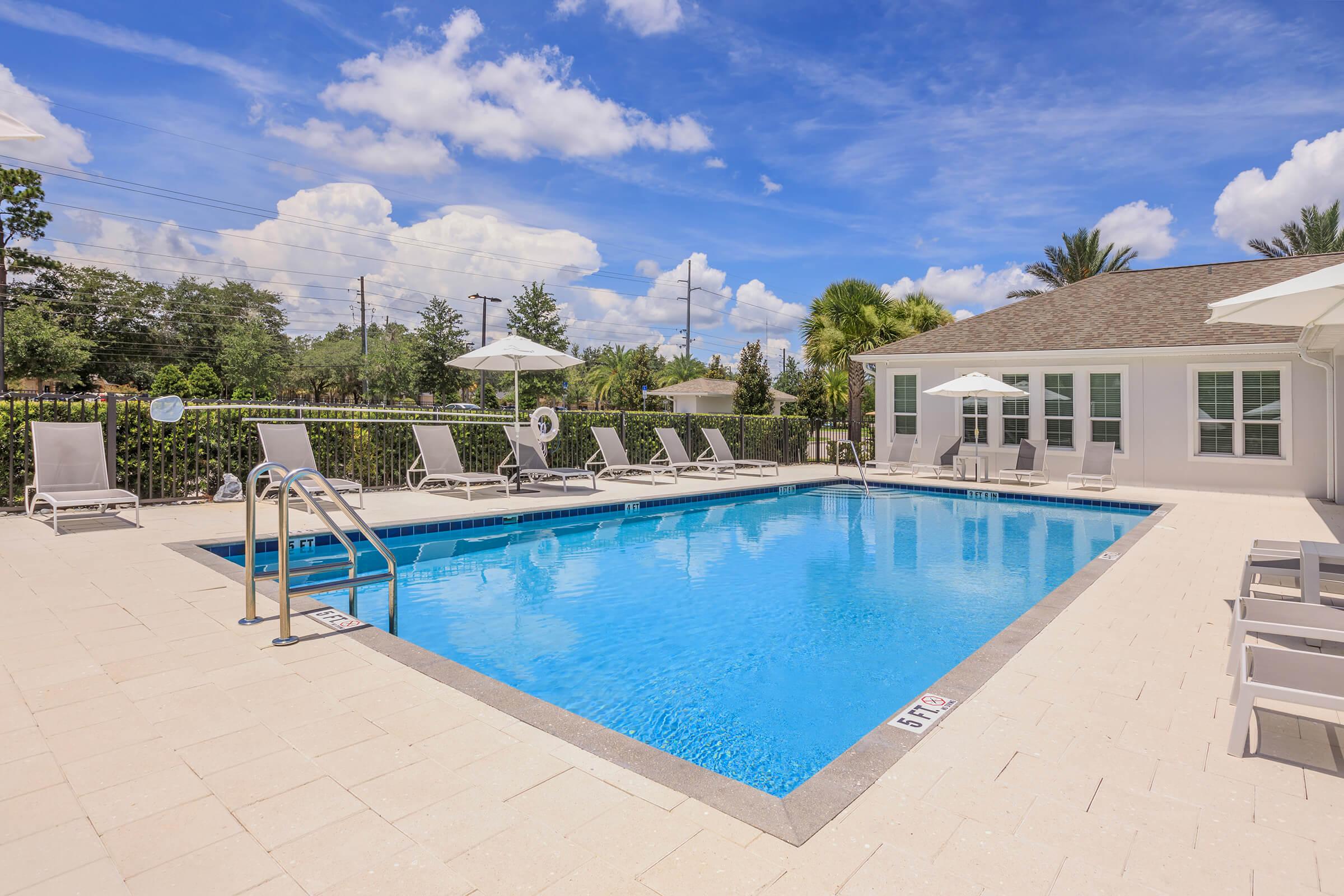 A clear, inviting swimming pool surrounded by lounge chairs and umbrellas, set against a backdrop of blue skies and fluffy white clouds. A modern building is visible to the right, with green trees in the background, creating a relaxing outdoor atmosphere.