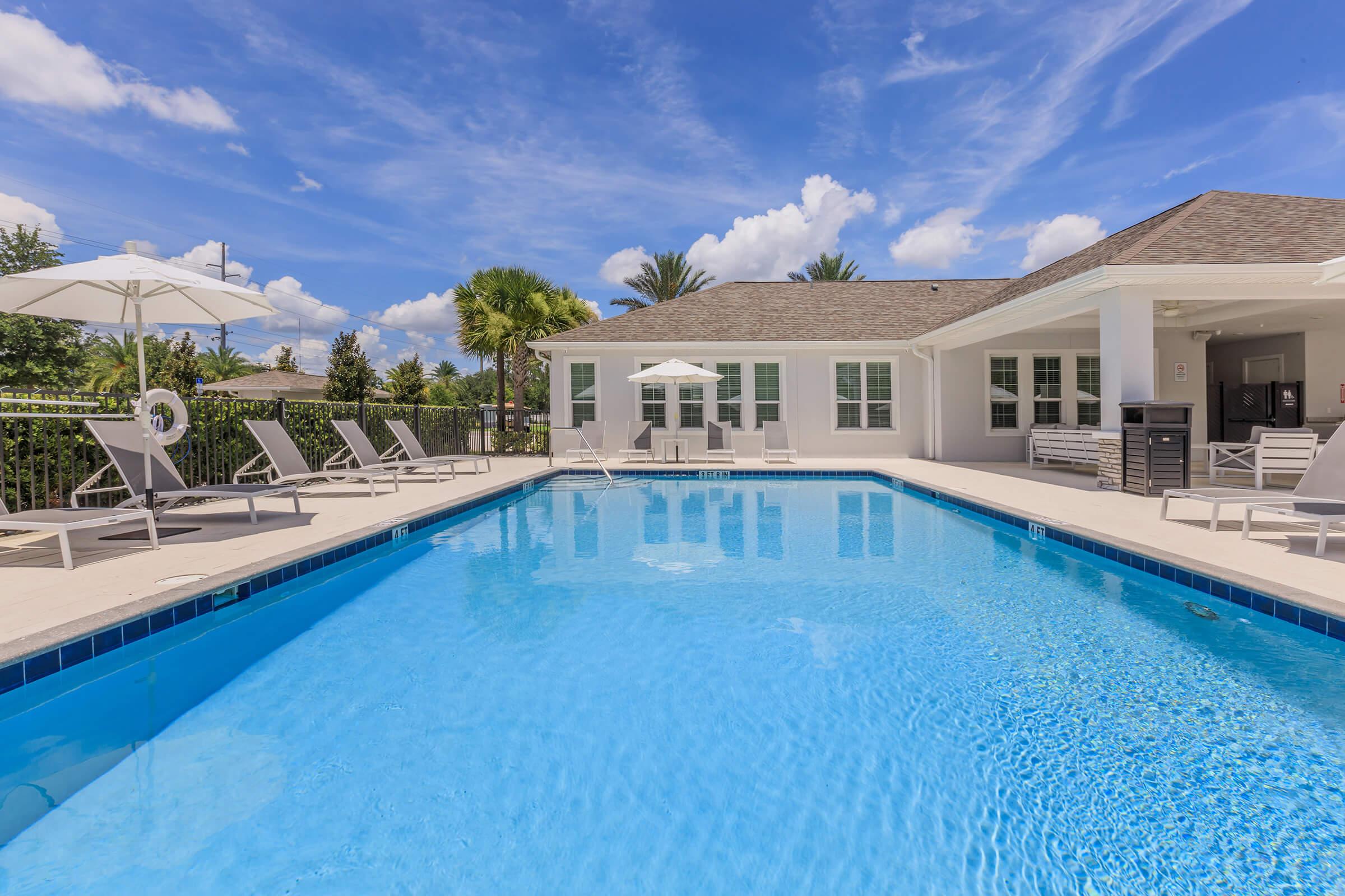 A bright outdoor pool surrounded by lounge chairs and umbrellas, with a modern house in the background. The sky is clear with fluffy white clouds, and palm trees are visible nearby, creating a tropical and inviting atmosphere.