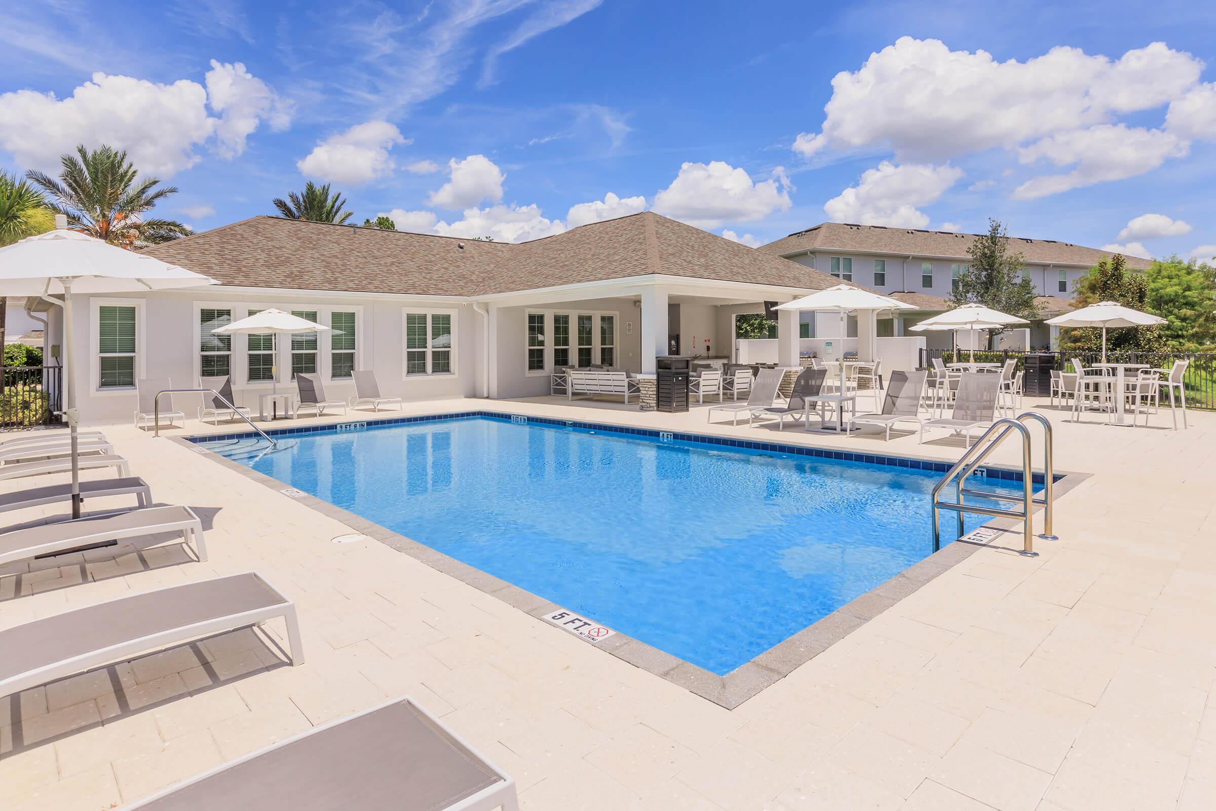 A serene outdoor pool area featuring a bright blue swimming pool surrounded by lounge chairs and umbrellas. A stylish building with large windows is visible in the background, and palm trees add a tropical feel to the scene. The sky is partly cloudy, creating a peaceful atmosphere.