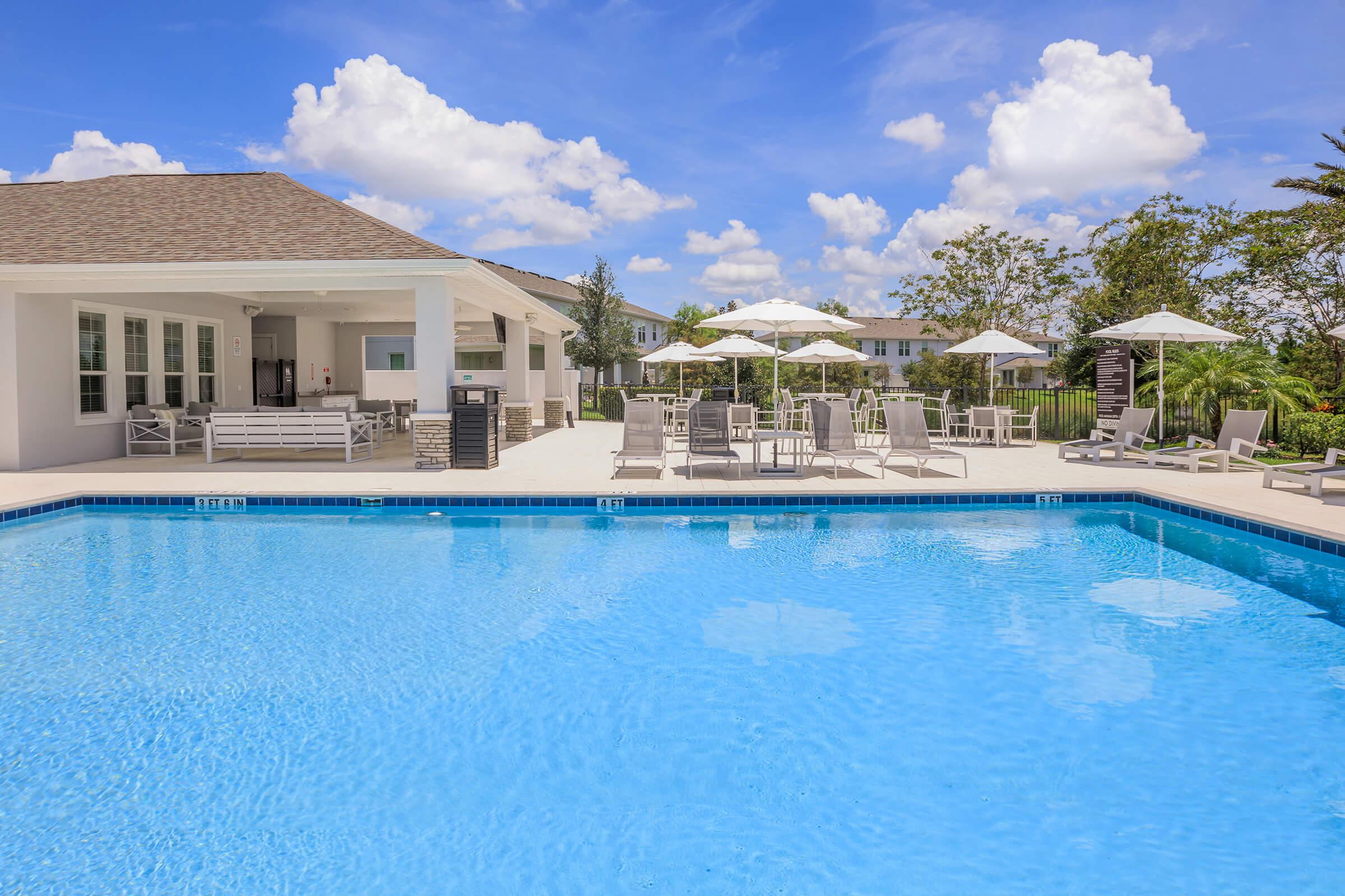 A swimming pool with clear blue water, surrounded by lounge chairs and umbrellas. In the background, a modern building with outdoor seating is visible under a bright blue sky filled with fluffy white clouds. Lush greenery is present around the pool area, creating a relaxed outdoor atmosphere.