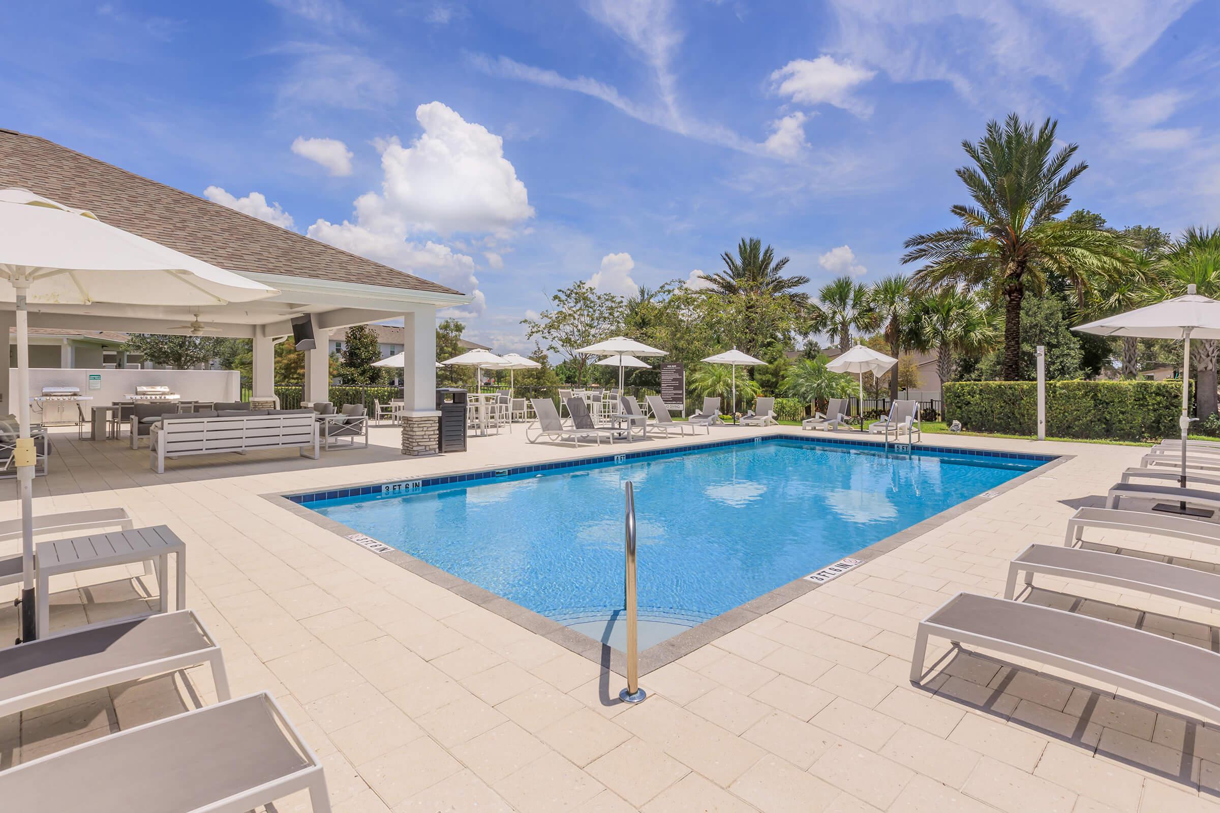 A sunny outdoor pool area featuring a clear blue pool surrounded by lounge chairs and umbrellas. In the background, lush palm trees can be seen, and a modern building provides shaded seating. The sky is bright with a few fluffy clouds, creating a relaxing vacation atmosphere.