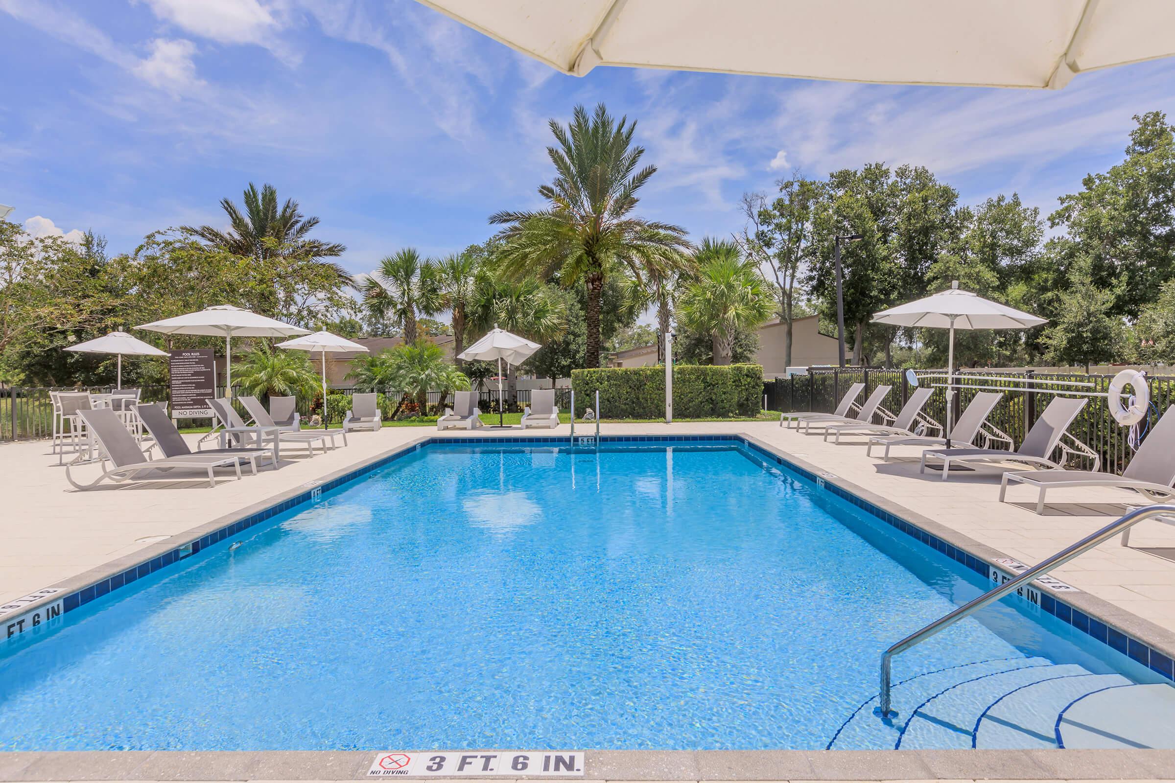 A clear blue swimming pool surrounded by lounge chairs and umbrellas. Lush palm trees and greenery are visible in the background, creating a tropical atmosphere. The pool area features a safety sign indicating the shallow end depth of 3 feet 6 inches.