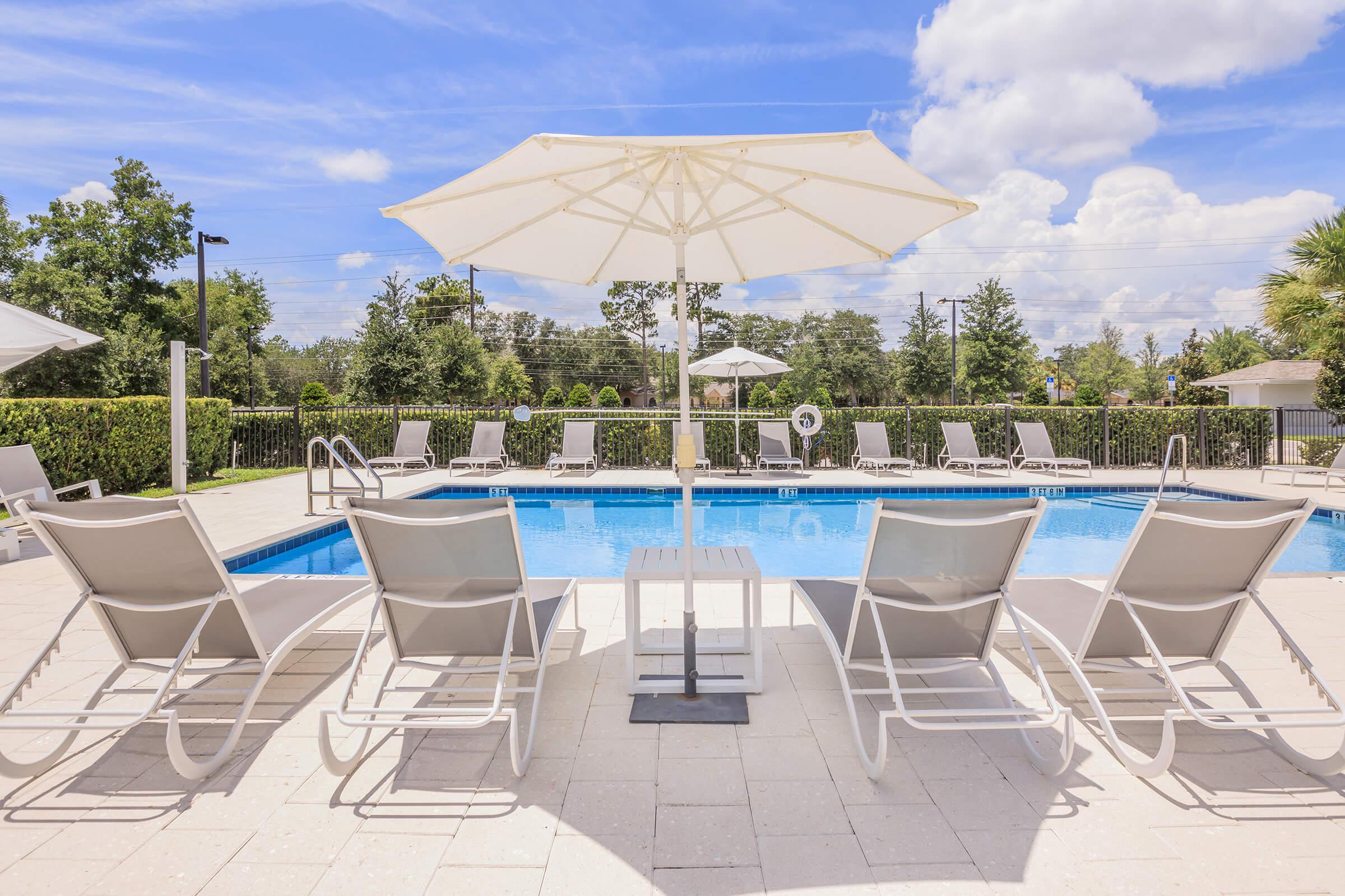 A serene outdoor pool surrounded by lounge chairs and an umbrella. Lush greenery and trees are visible in the background, with a clear blue sky and fluffy white clouds above. The pool deck features light-colored tiles, creating a relaxing atmosphere for leisure and sunbathing.