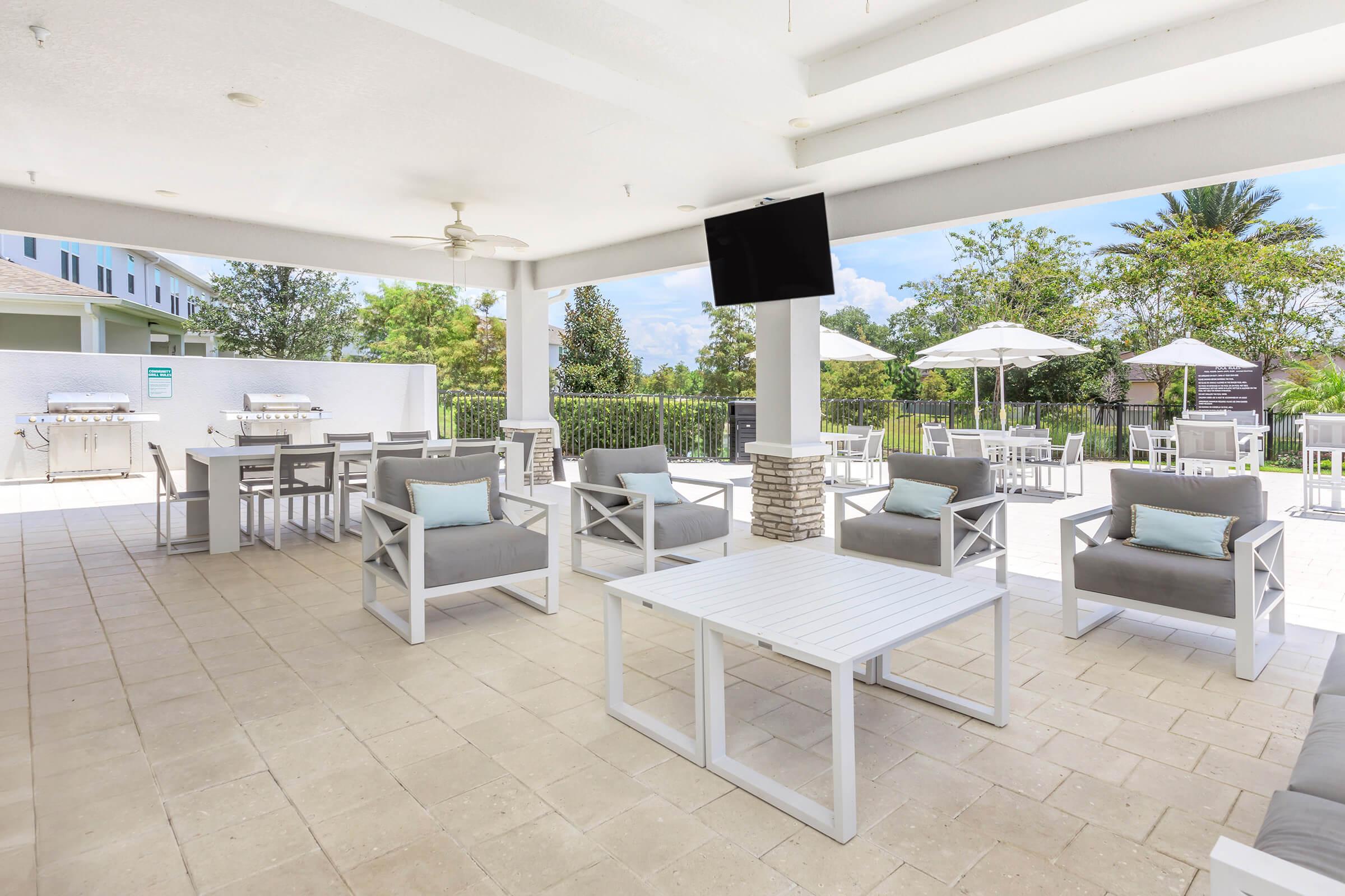 Open outdoor lounge area featuring white furniture, including chairs and tables. Gray cushioned armchairs are arranged around a white table. In the background, there are barbecue grills, umbrella-covered tables, and lush greenery. A television mounted on the wall adds to the recreational setting.