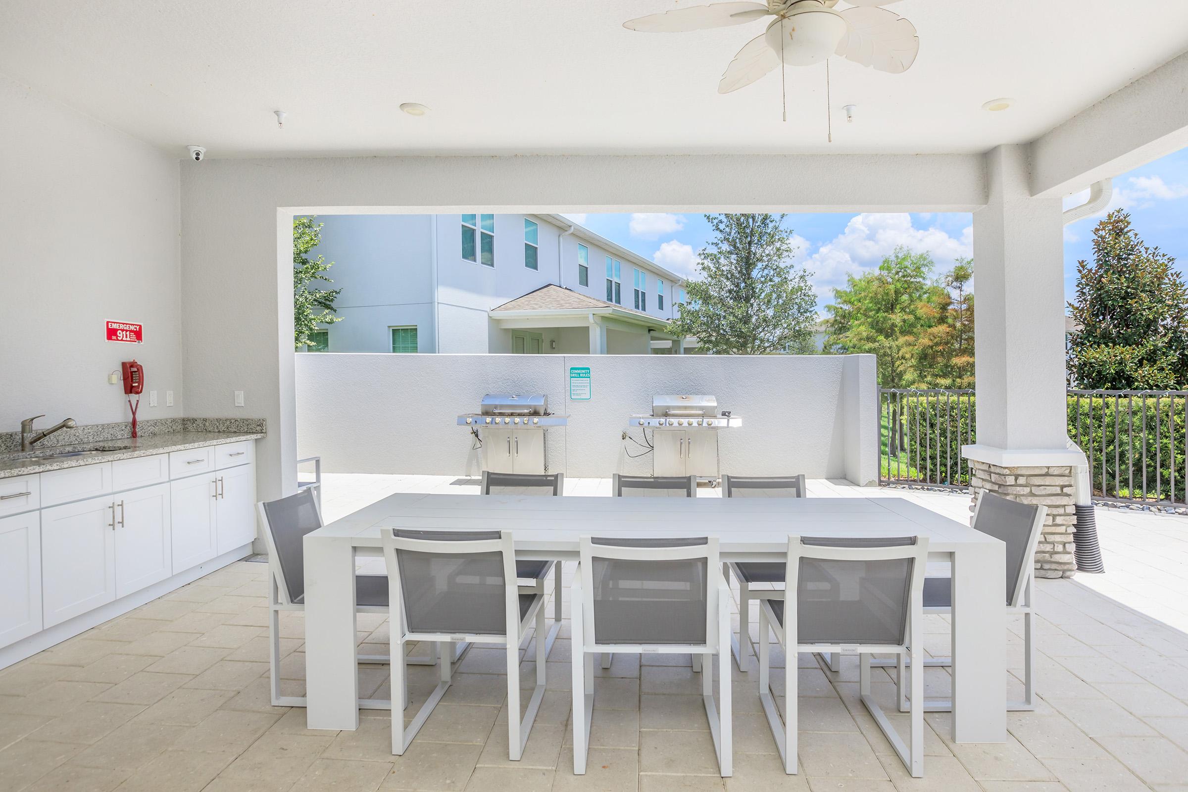 Outdoor dining area with a long white table and gray chairs, surrounded by walls and greenery. Two stainless steel grills are visible in the background. The space is well-lit with a ceiling fan overhead, offering a modern and inviting atmosphere for gatherings.