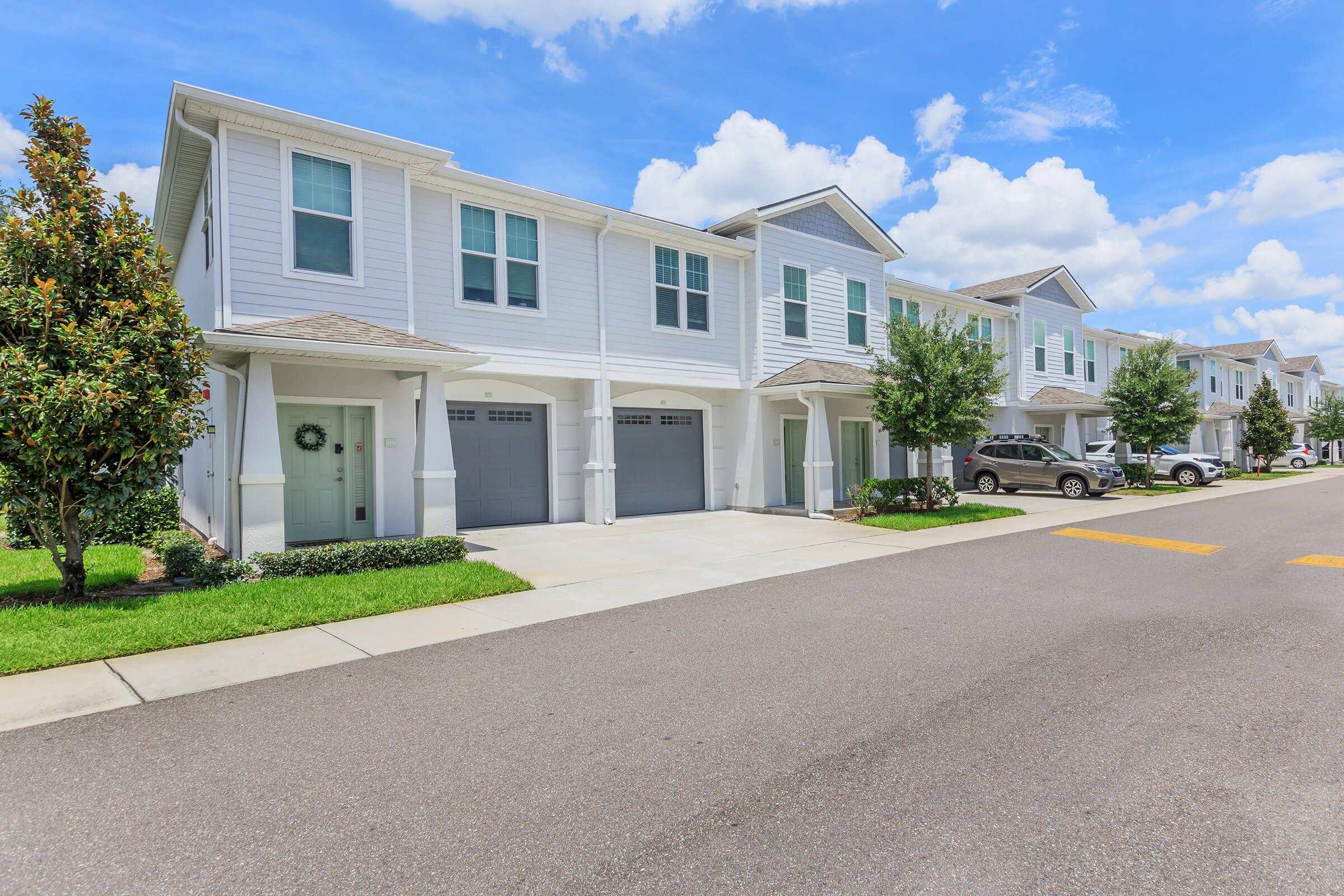 A suburban street featuring a row of modern two-story residential buildings with garages. Each unit has a light-colored exterior and a small porch. Lush green lawns and trees line the sidewalk, while blue skies and fluffy white clouds create a bright and inviting atmosphere.